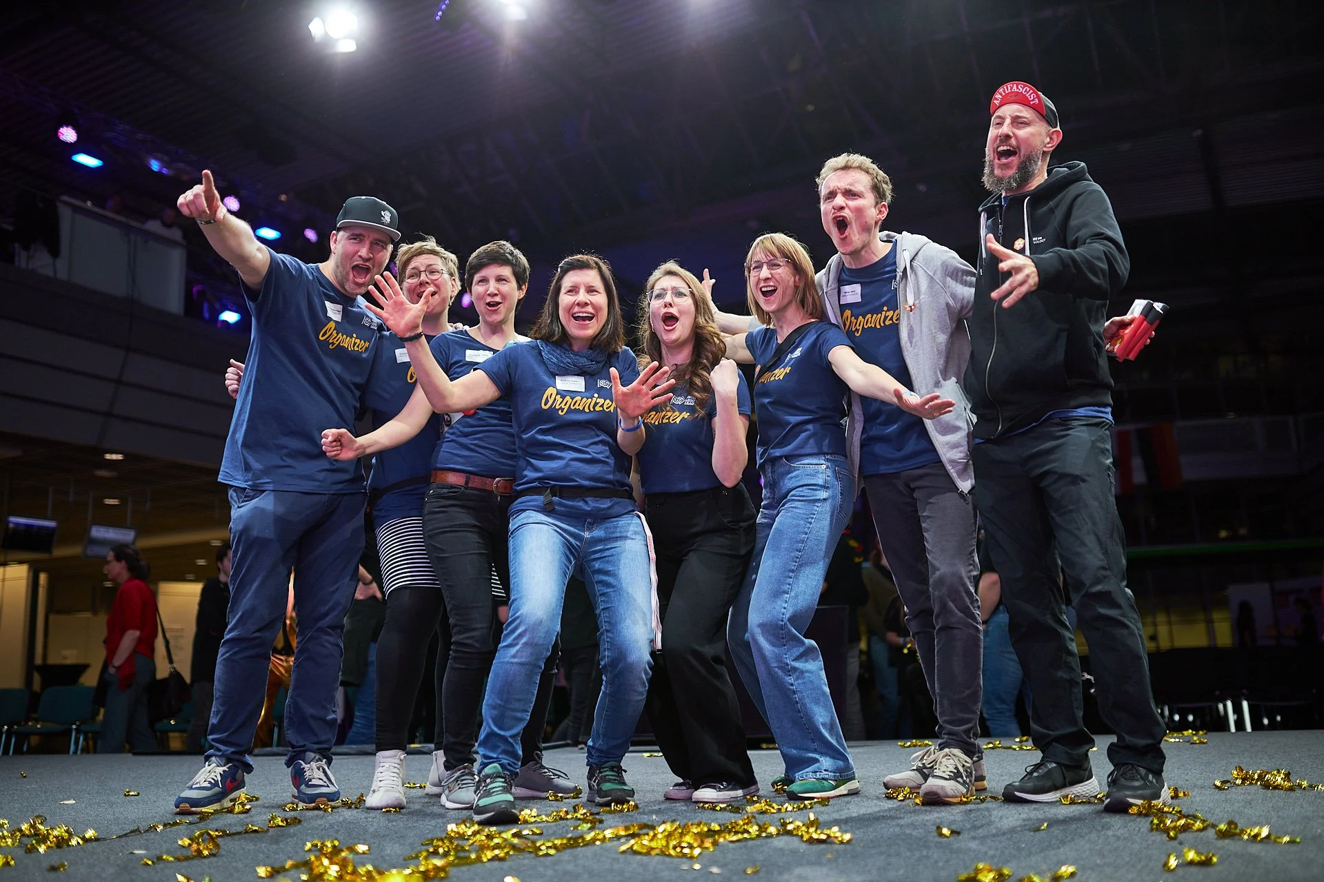 Eine Gruppe von Menschen auf einer Bühne, die jubeln, tanzen und sich umarmen, während sie in blauen T-Shirts mit der Aufschrift 'Organizer' tragen, um feiern oder einen Erfolg zu feiern.