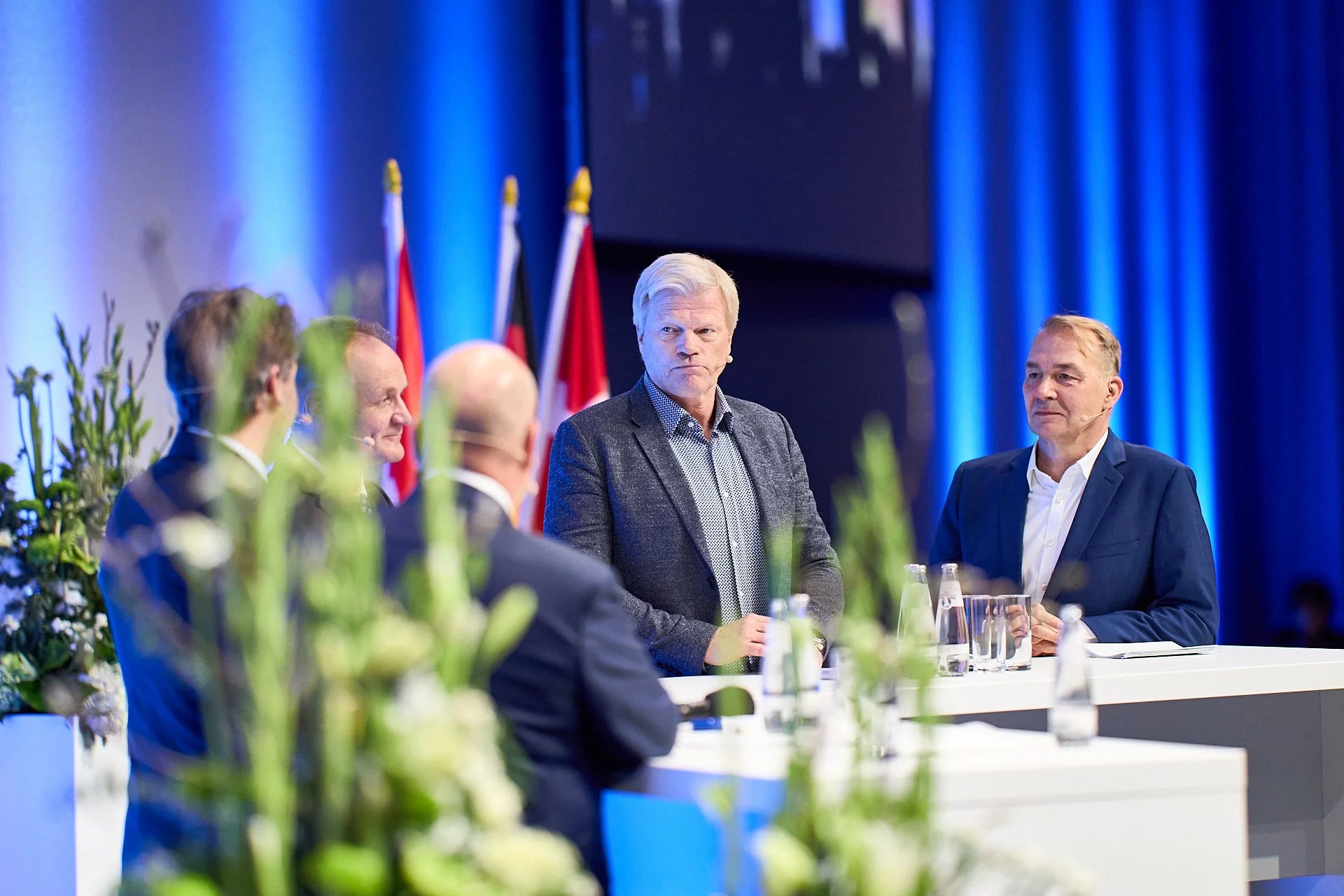 Mehrere Männer sitzen an einem Konferenztisch auf einer Bühne mit blauen Vorhängen und Flaggen im Hintergrund, darunter Deutschland und Norwegen.