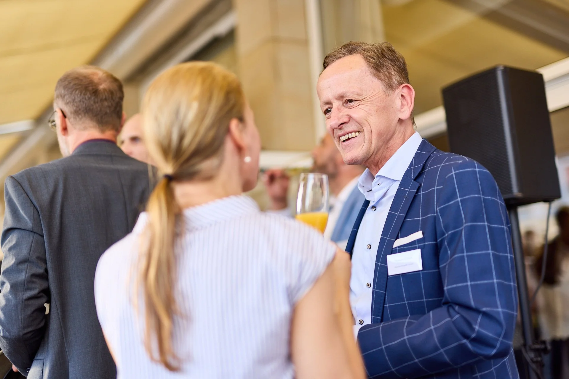 Zwei Personen im Gespräch bei einer formellen Veranstaltung, eine Frau mit blonden Haaren und ein Mann in einem blauen karierten Anzug, der eine orangefarbene Getränk in der Hand hält.