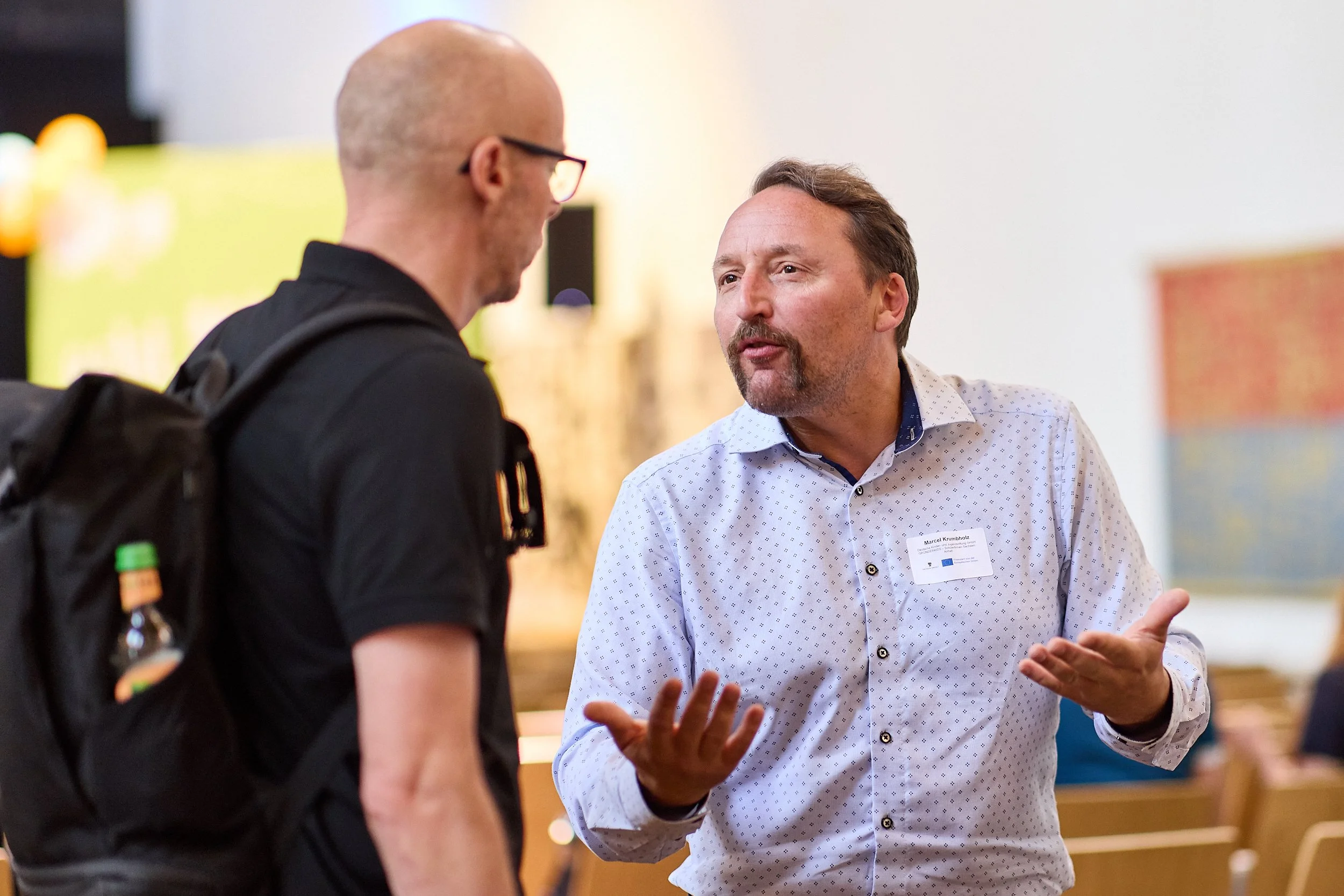 Zwei Männer im Gespräch bei einer Veranstaltung, einer erklärt etwas und der andere hört aufmerksam zu, im Hintergrund Sitzbänke.