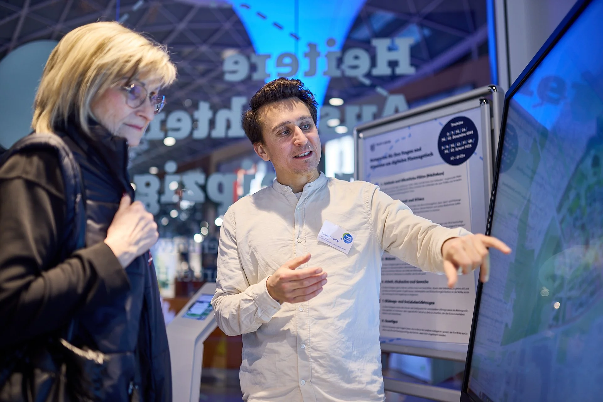 Ein Mann zeigt auf eine große Karte oder einen Bildschirm, während eine Frau mit Brille ihm zuhört, bei einer Informationsveranstaltung.