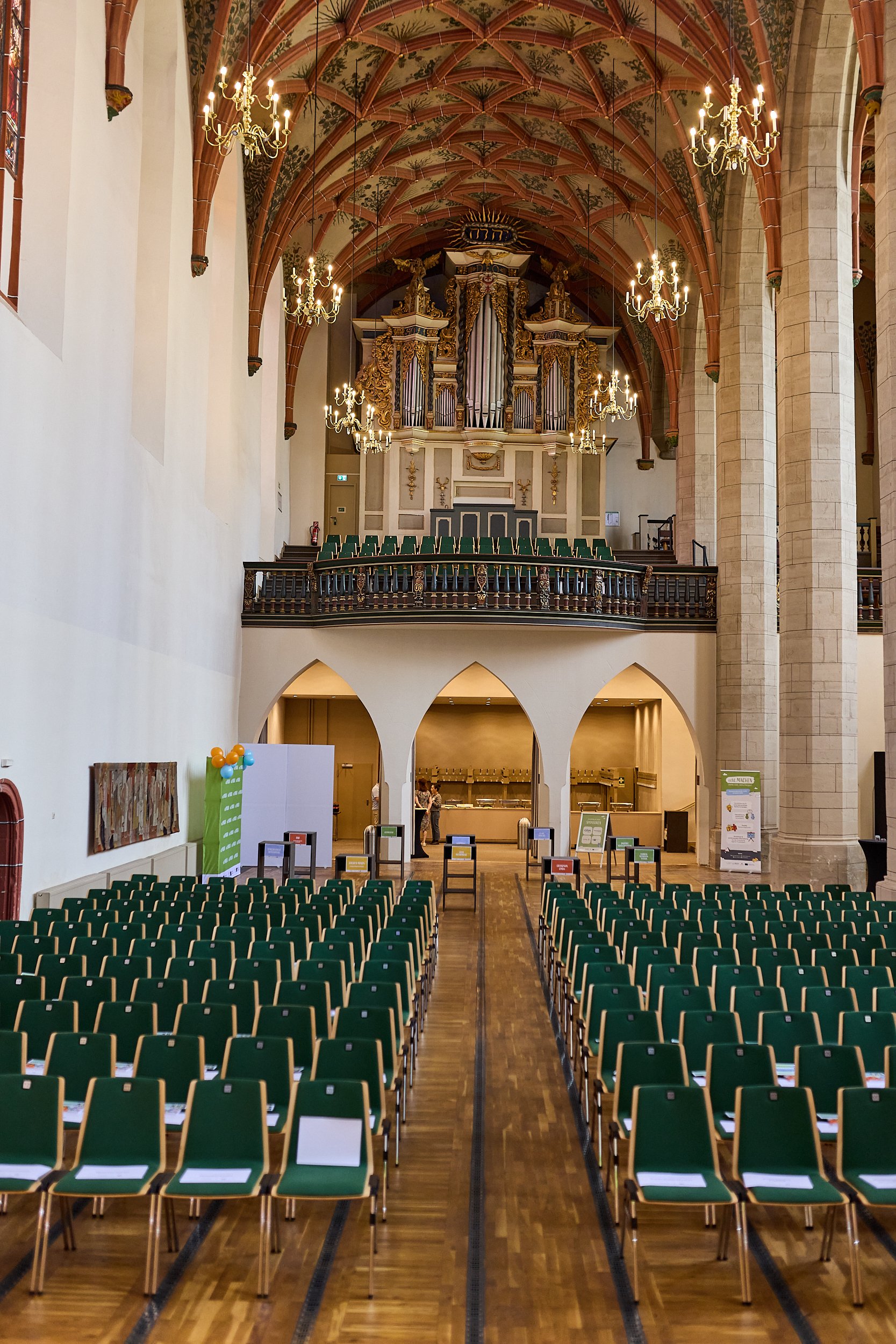 Ein großer, prunkvoller Saal mit einem high ceiling, vielen grünen Stühlen in Reihen, einem Obergeschoss mit einer Orgel und einem Christuskreuz an der Decke. Es gibt auch mehrere Kronleuchter und einen Empfangstresen im Hintergrund.