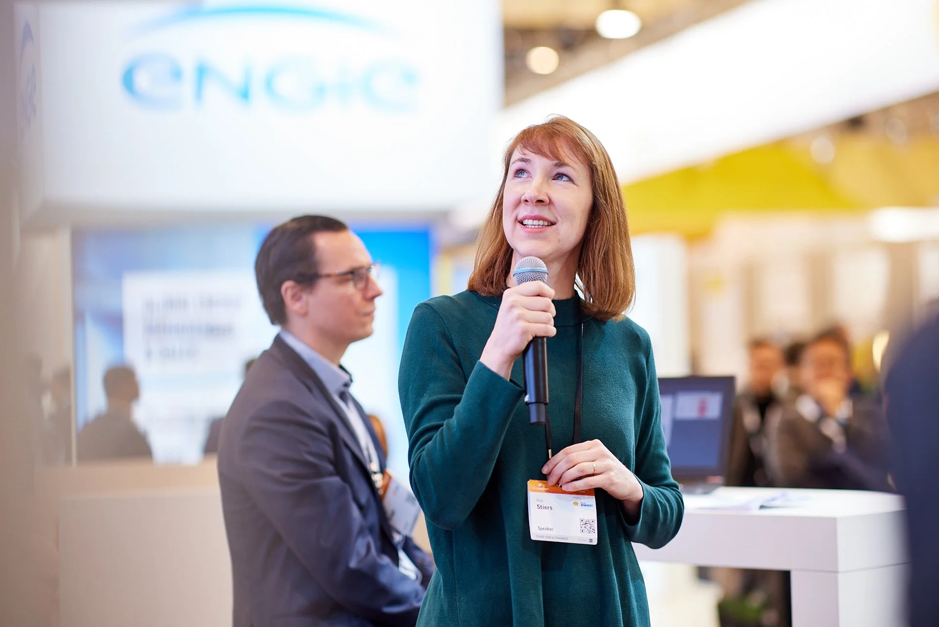 Eine Frau spricht mit einem Mikrofon auf einer Konferenz, im Hintergrund sind andere Personen und ein Bildschirm mit dem Logo »CNCG« sichtbar.