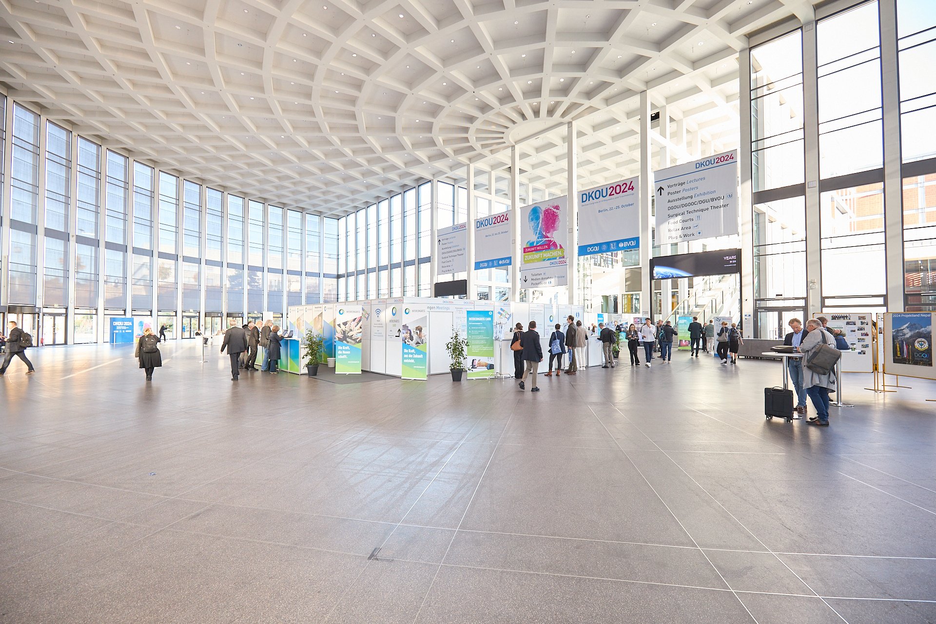 Innenansicht einer großen, modernen Konferenzhalle mit hoher, gewölbter Decke und viel natürlichem Licht durch große Fensterflächen. Mehrere Menschen sind an Messeständen und beim Austausch. Es gibt Ausstellungstafeln, Banner und digitale Bildschirme