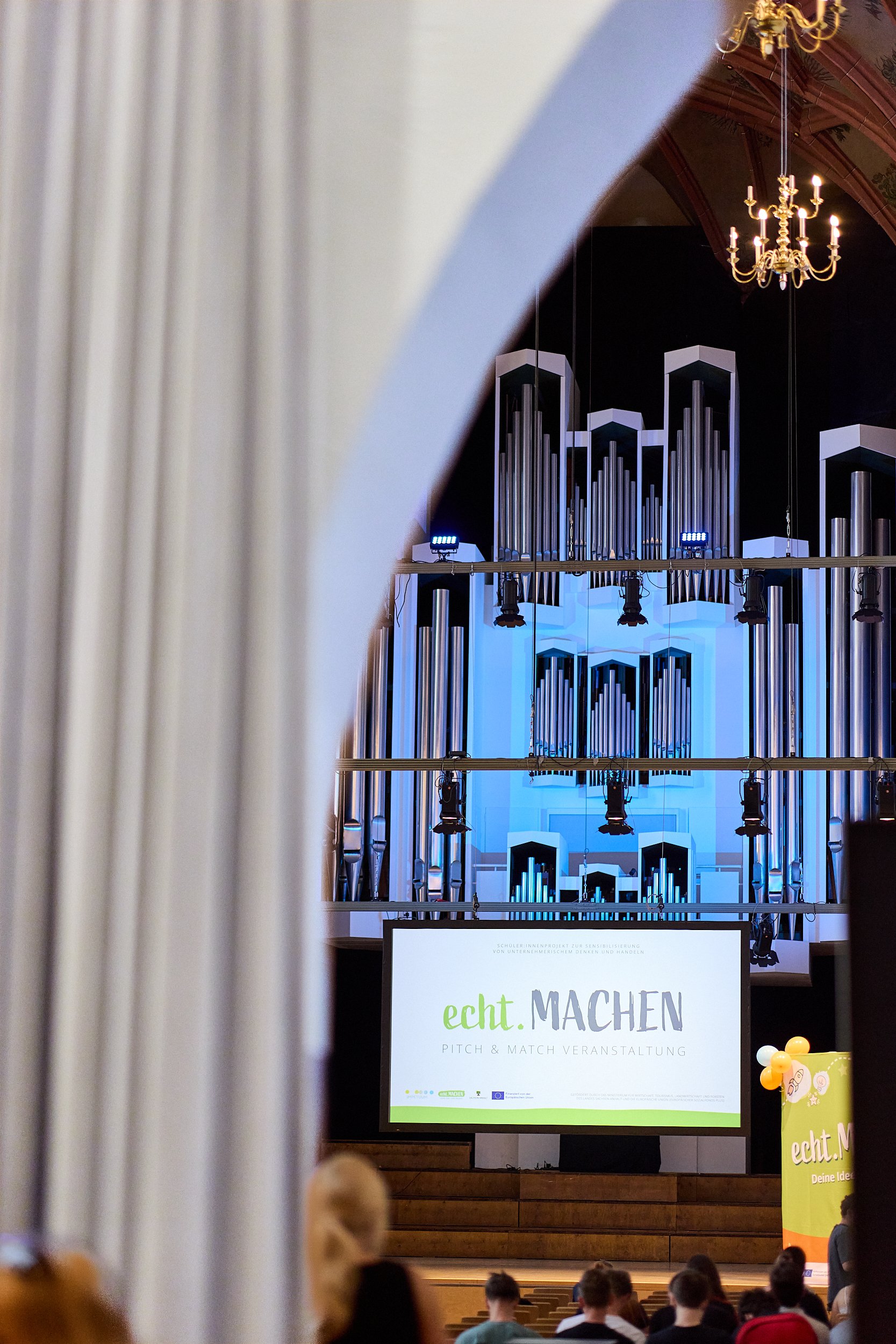 Blick auf eine Bühne mit einem großen digitalen Bildschirm, auf dem 'echt. MACHEN' steht, und im Hintergrund eine große Orgel. Es sitzen mehrere Menschen im Saal vor der Bühne.