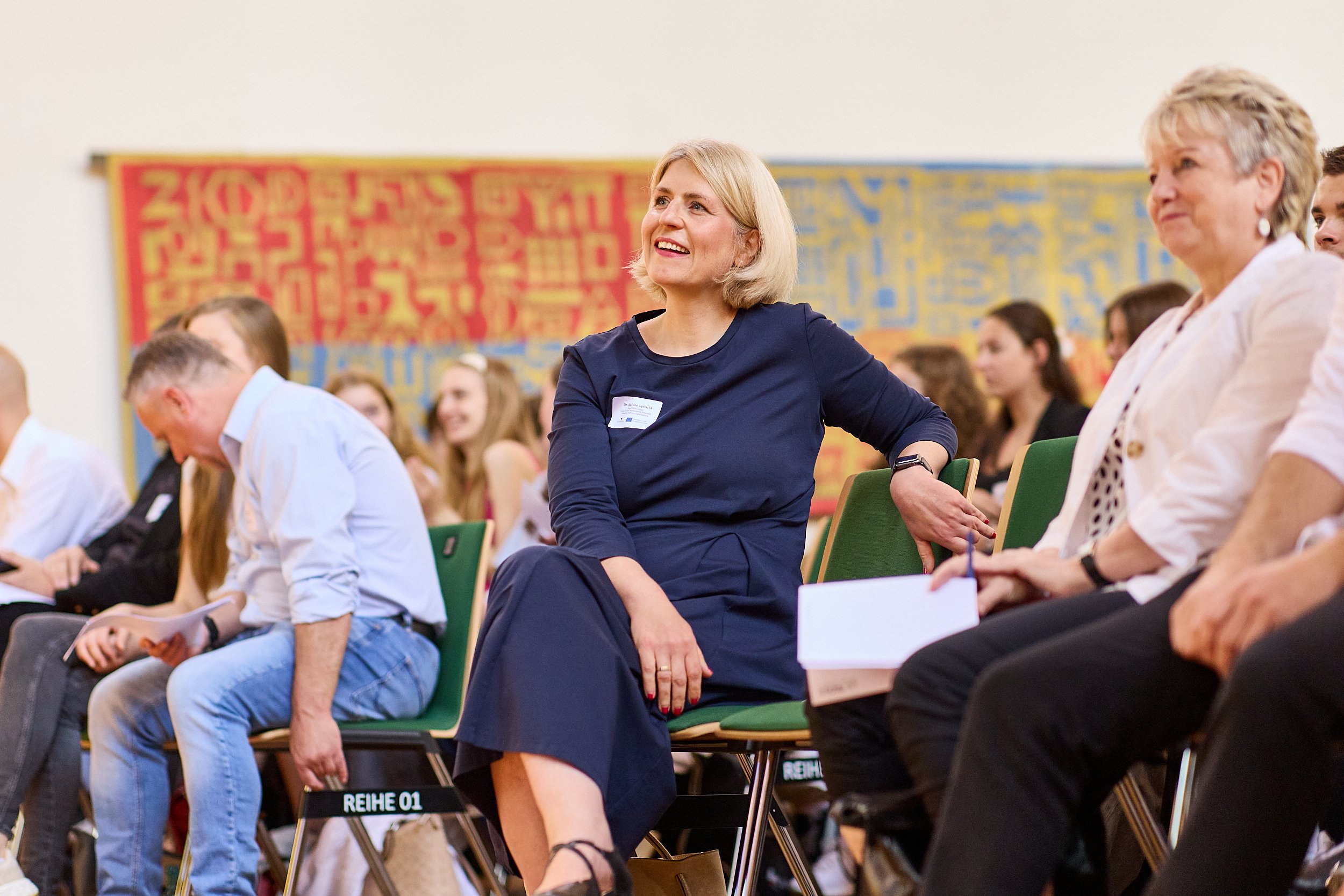 Frau in dunklem Kleid sitzt bei einer Veranstaltung, lacht und schaut nach vorne.