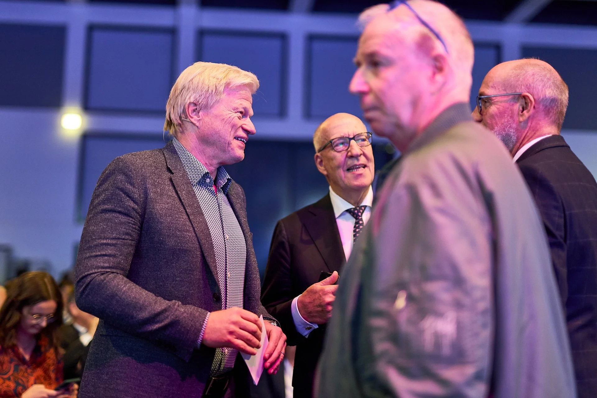 Vier ältere Männer in formeller Kleidung unterhalten sich bei einer Veranstaltung, im Hintergrund sind weitere Personen zu sehen.