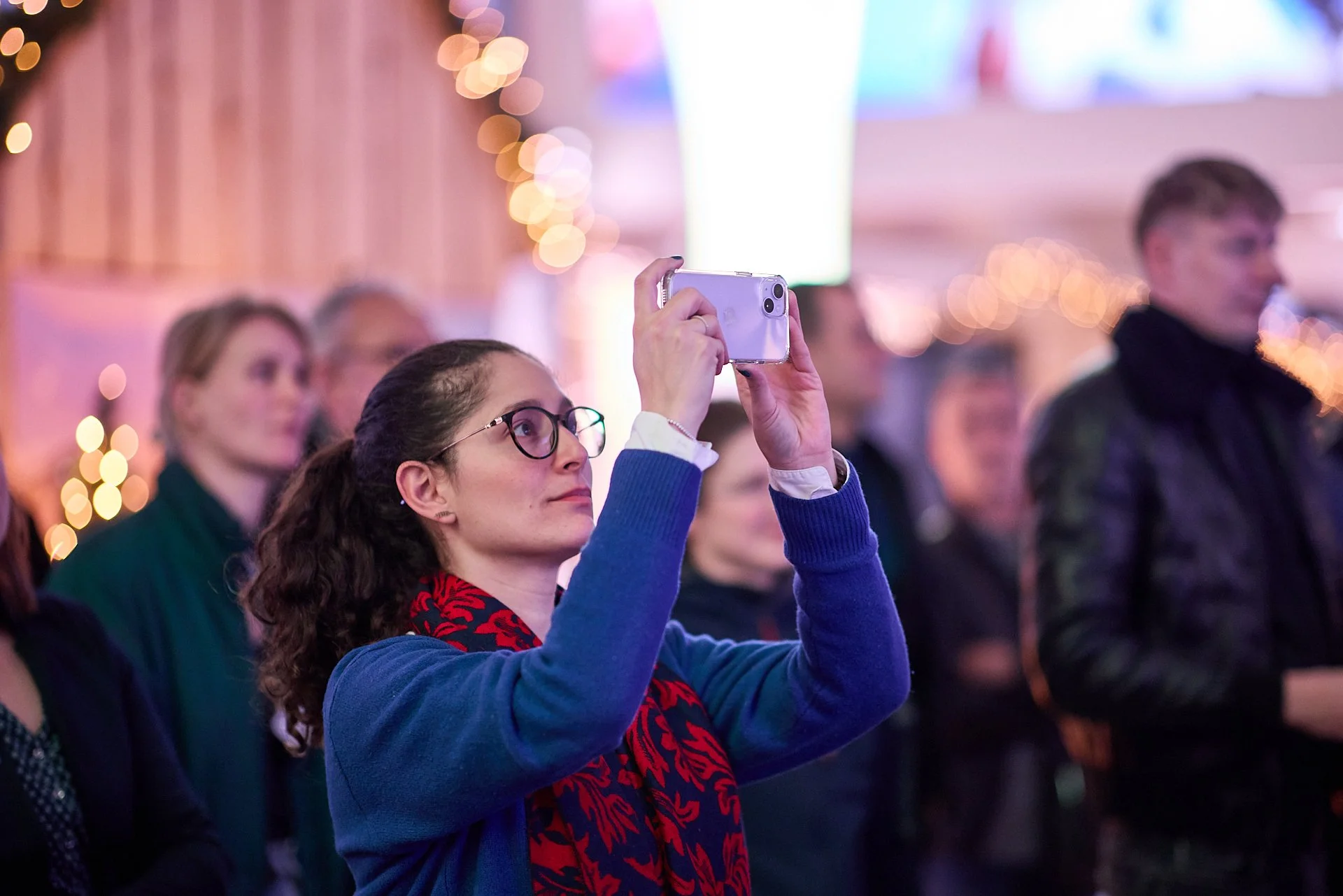 Eine Frau mit Brille und dunklen, lockigen Haaren fotografiert mit einem Smartphone bei einer Veranstaltung, umgeben von anderen Gästen.