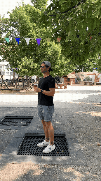 Ein junger Mann steht auf einem Gitter im Freien, trägt schwarzen T-Shirt, graue Shorts, weißes Cap, Sonnenbrille und hält ein Getränk in der Hand. Im Hintergrund sind Bäume, ein Spielplatz und bunte Fahnen.