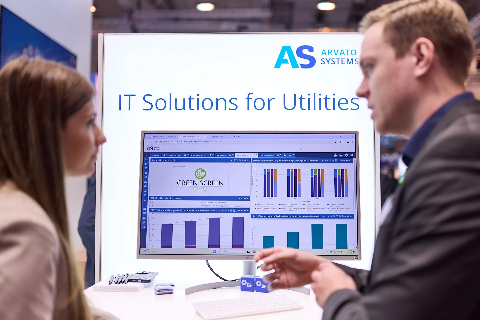Zwei Personen im Gespräch vor einem Stand mit einem großen Bildschirm, der Diagramme und Grafiken zeigt, und einem weißen Hintergrund mit blauer Schrift, die 'IT Solutions for Utilities' und das Logo 'AS Arvato Systems' trägt.