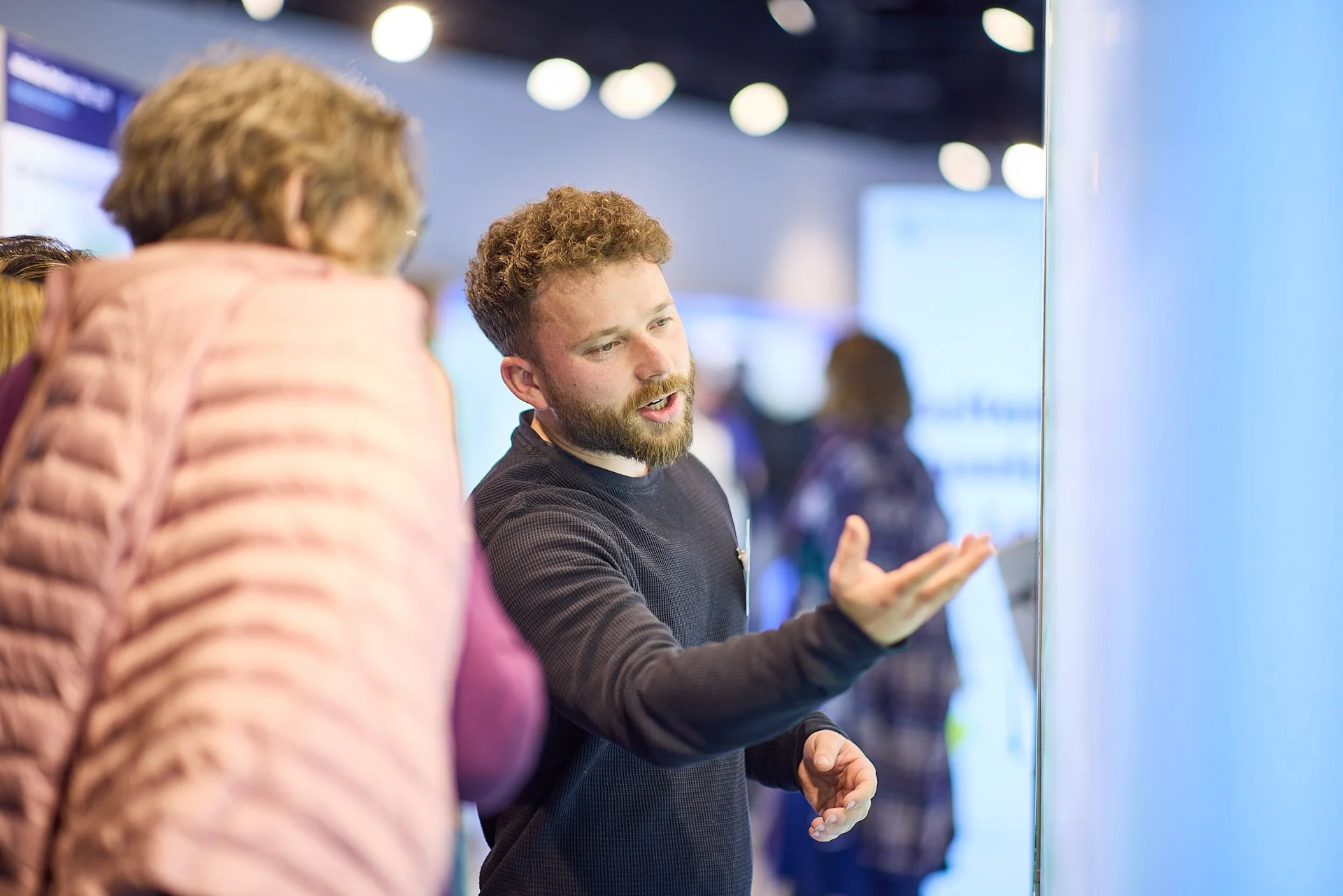 Zwei Menschen sprechen auf einer Messe oder Konferenz, die Person auf der rechten Seite erklärt etwas, während die Person auf der linken Seite zuhört.