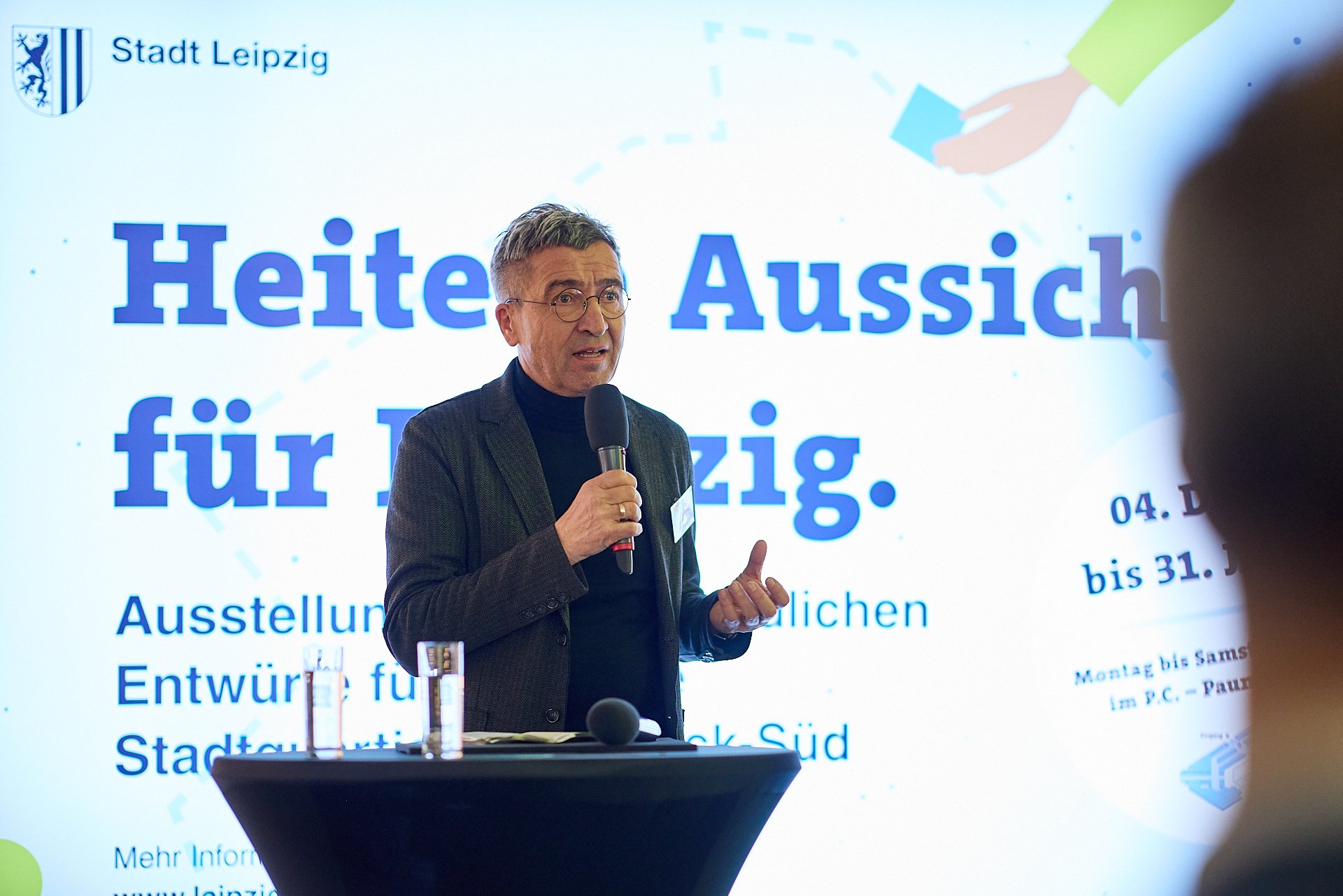 Ein Mann spricht bei einem Vortrag vor einer große Bildschirmprojektion, die den Text 'Heiter Aussichten für Leipzig' zeigt, mit Wasserflaschen auf einem Stehtisch vor ihm.