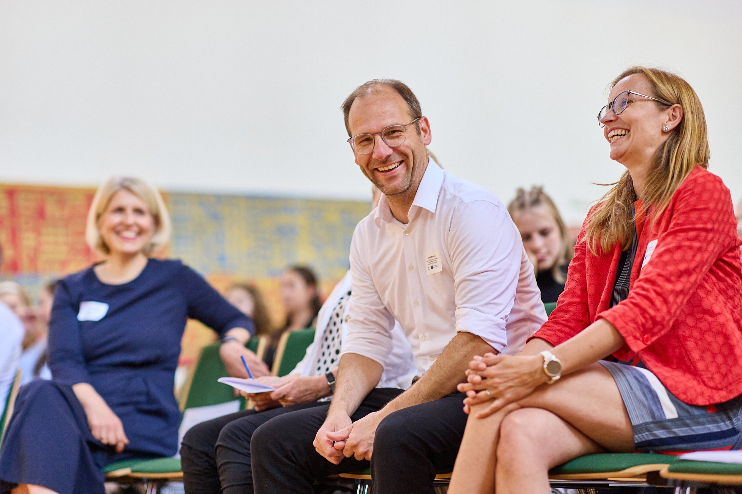 Menschen lachen und unterhalten sich bei einem Treffen in einem Raum mit grünen Stühlen und einer bunten Wand im Hintergrund.