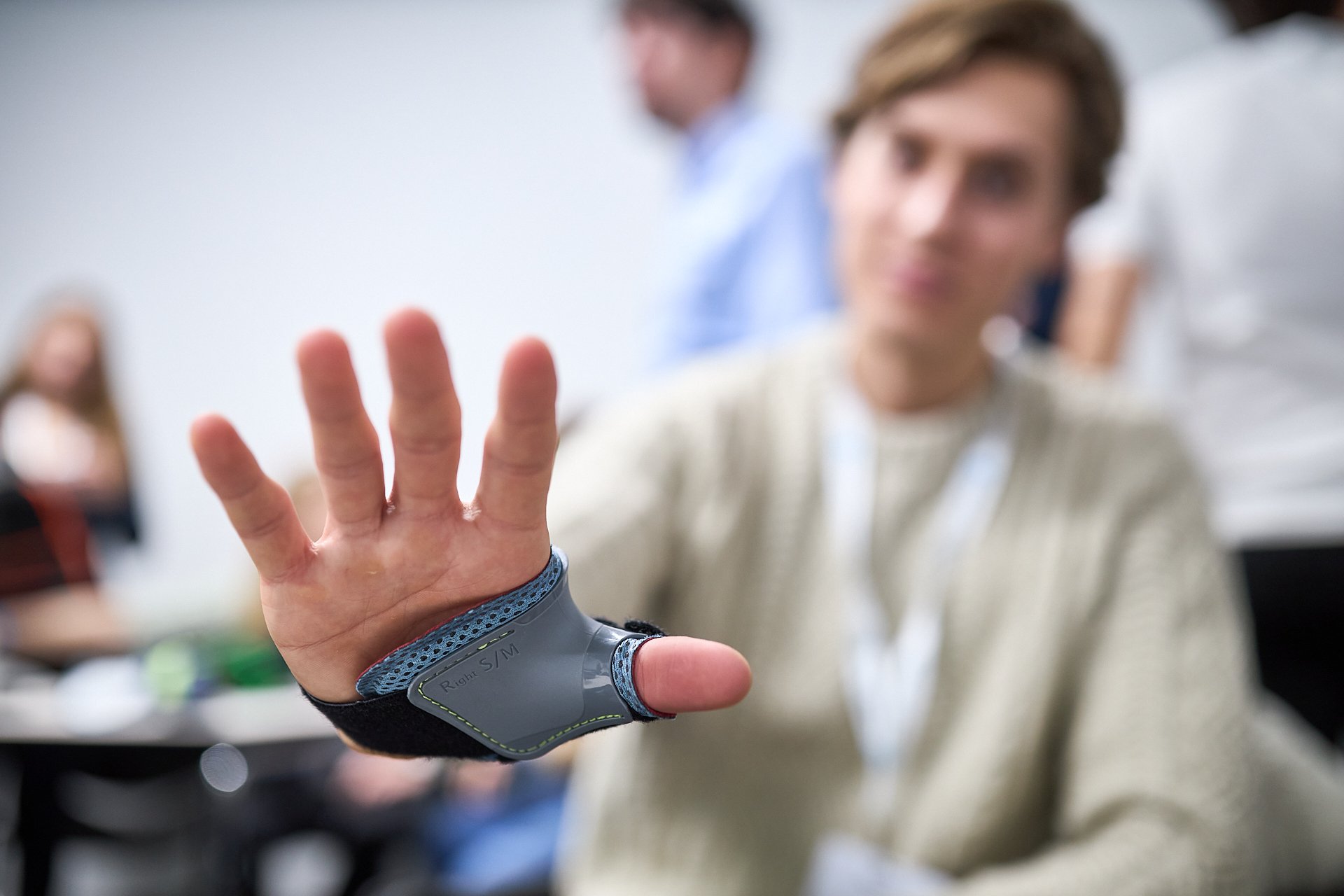 Ein Mann zeigt seine Hand mit einer Prothese am Arm, die einen elektronischen Handprotheseersatz darstellt, während er in einem Raum mit mehreren Personen sitzt.