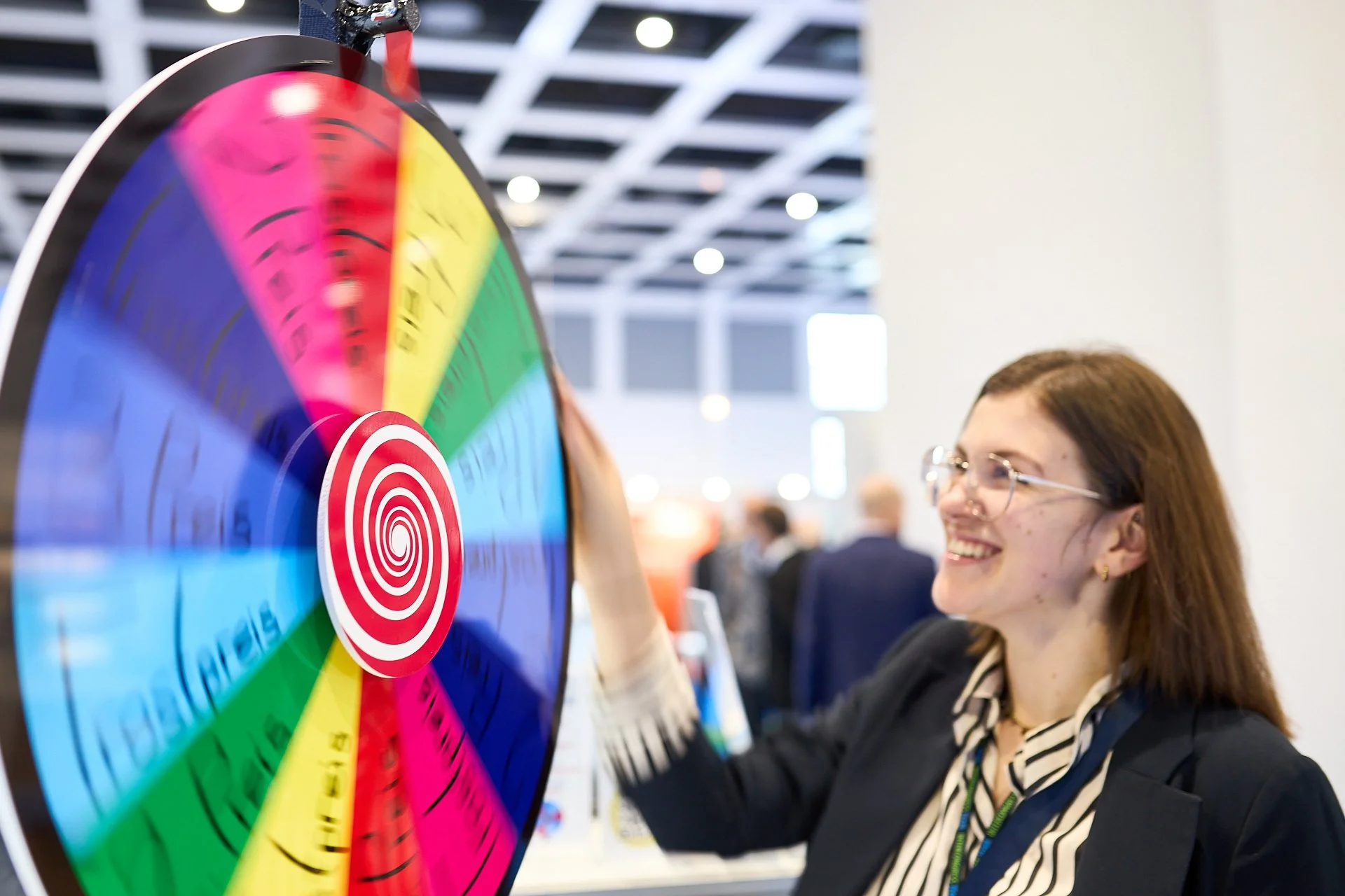 Eine lachende Frau mit Brille und dunklem Haar dreht an einem bunten Glücksrad bei einer Veranstaltung im Innenbereich.