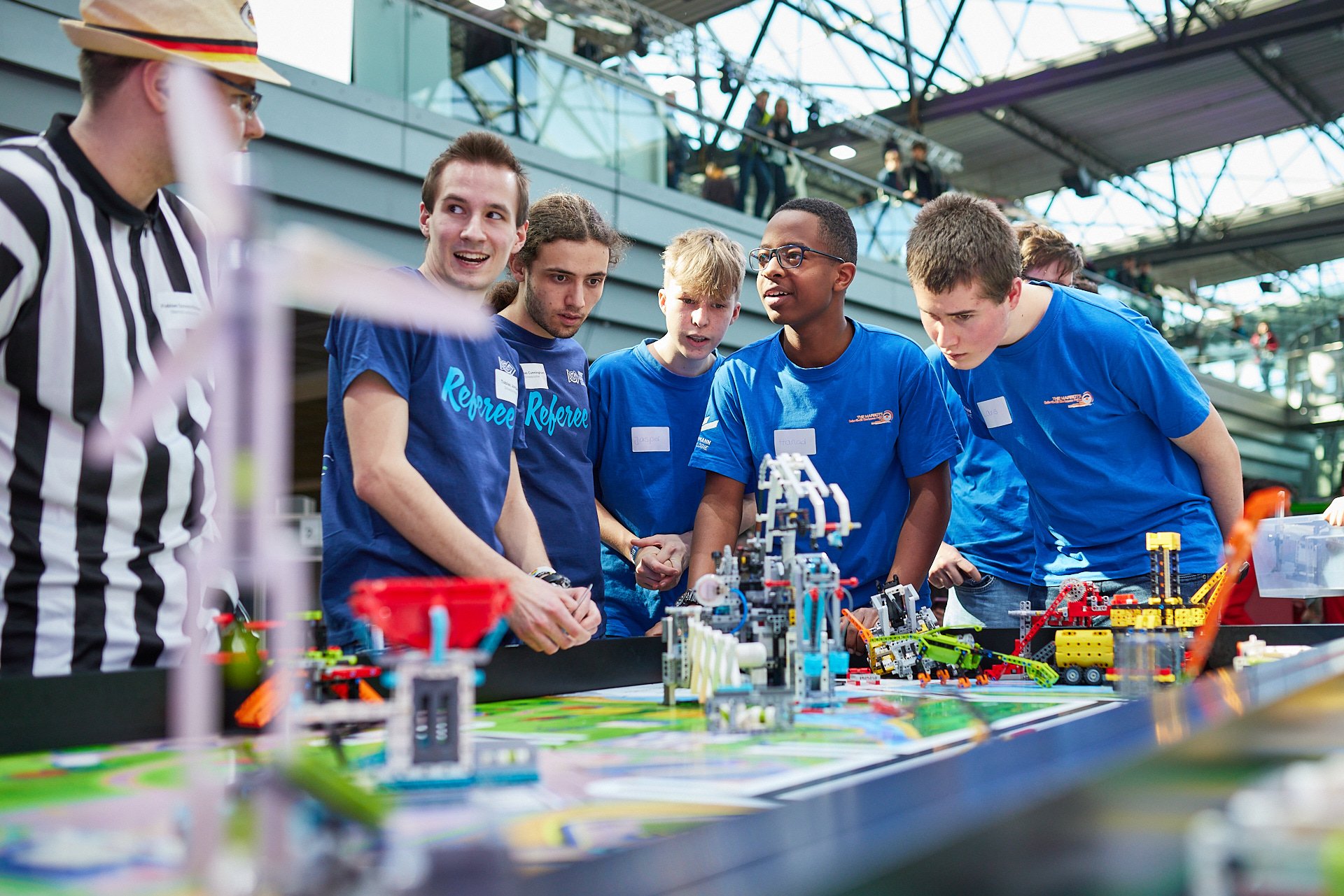Jugendliche beim Robotikwettbewerb in einer modernen Halle, umgeben von Robotern und Modelllandschaften, elektrisches Licht und Leute im Hintergrund.