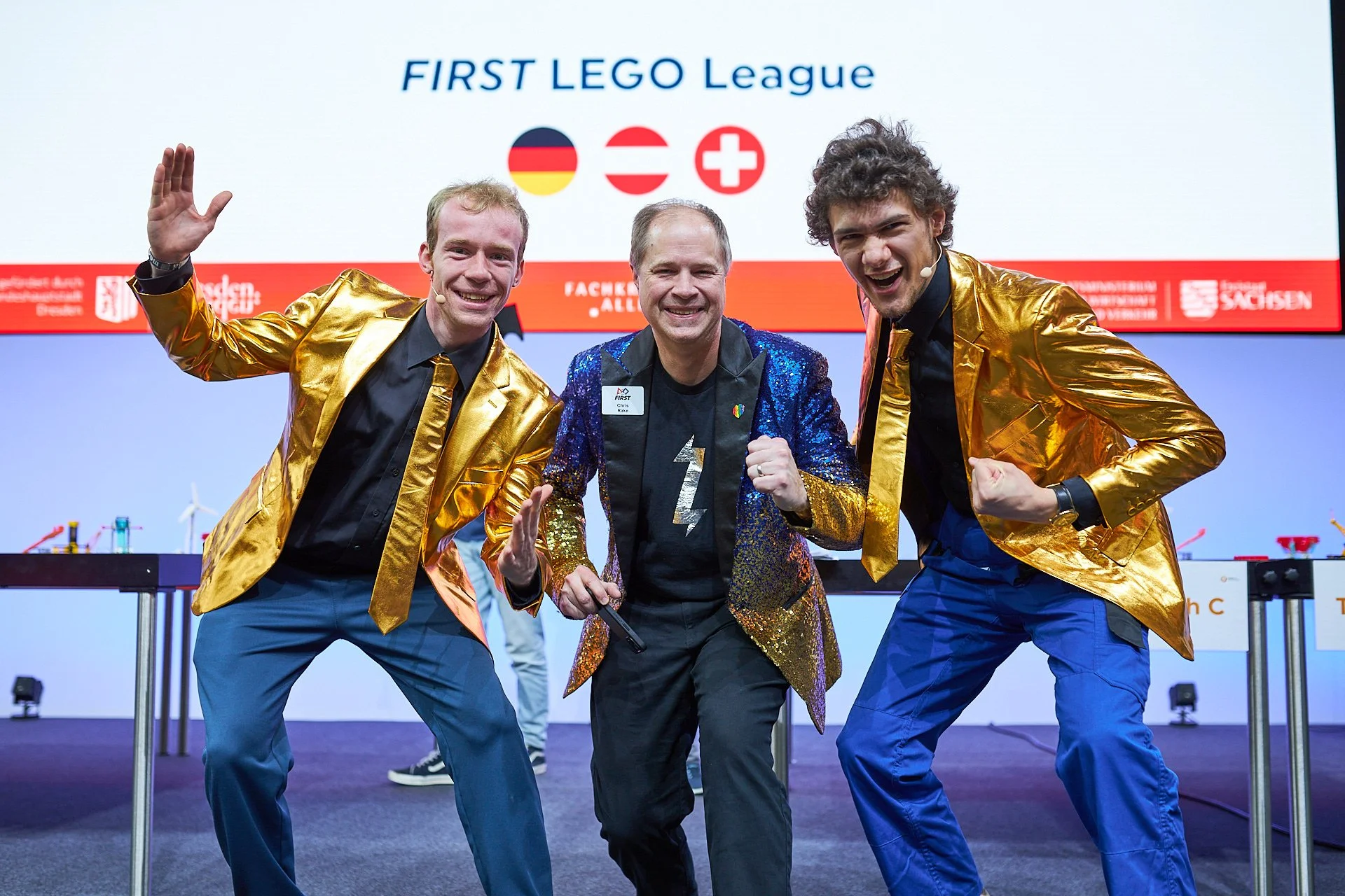 Drei Männer in goldenen und blauen Jacken feiern auf einer Bühne bei der FIRST LEGO League, vor einem Bildschirm mit Deutschland, Österreich und Schweiz Flaggen.