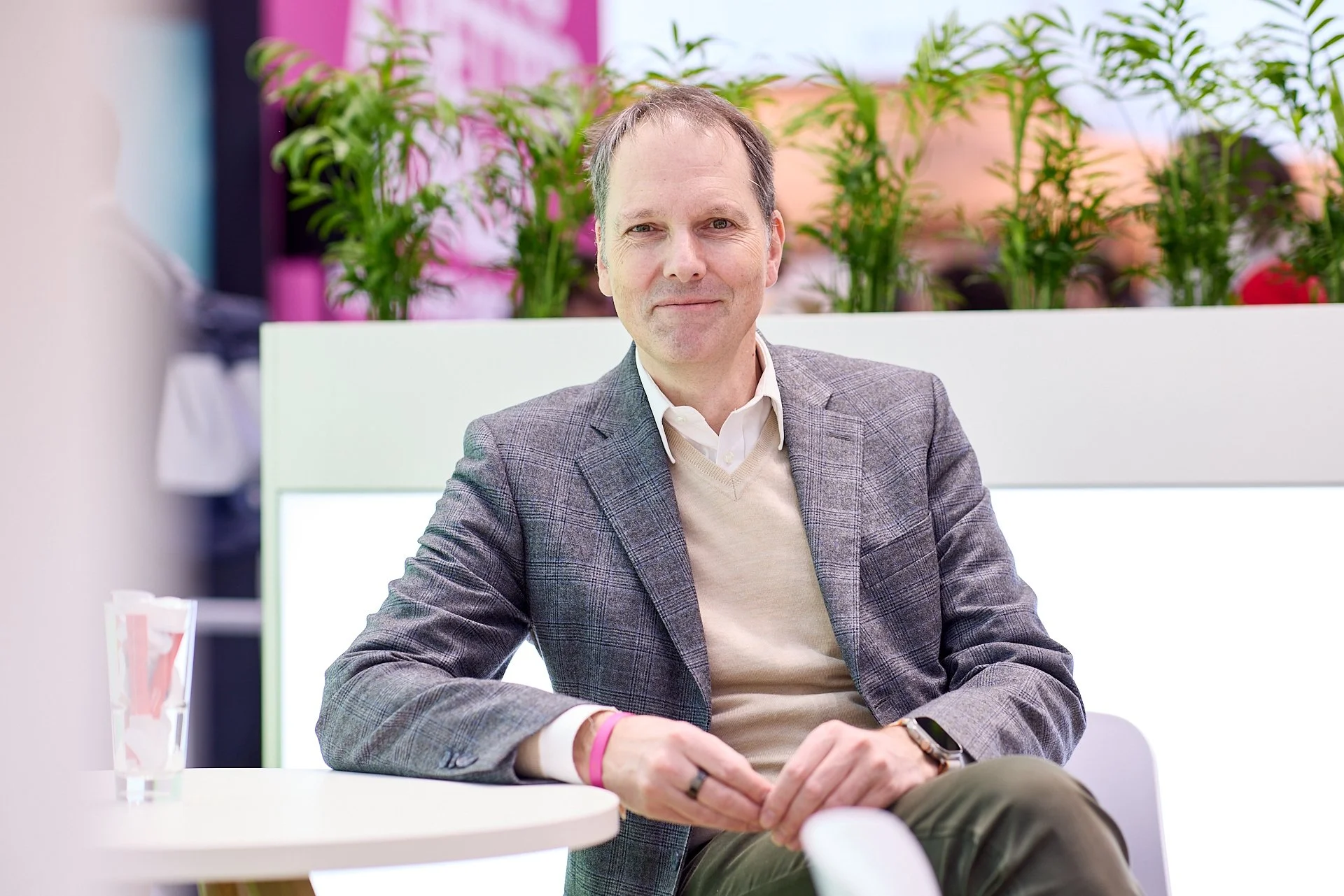 Ein Mann sitzt an einem weißen Tisch, trägt einen grauen Anzug und schaut in die Kamera, im Hintergrund sind grüne Pflanzen.