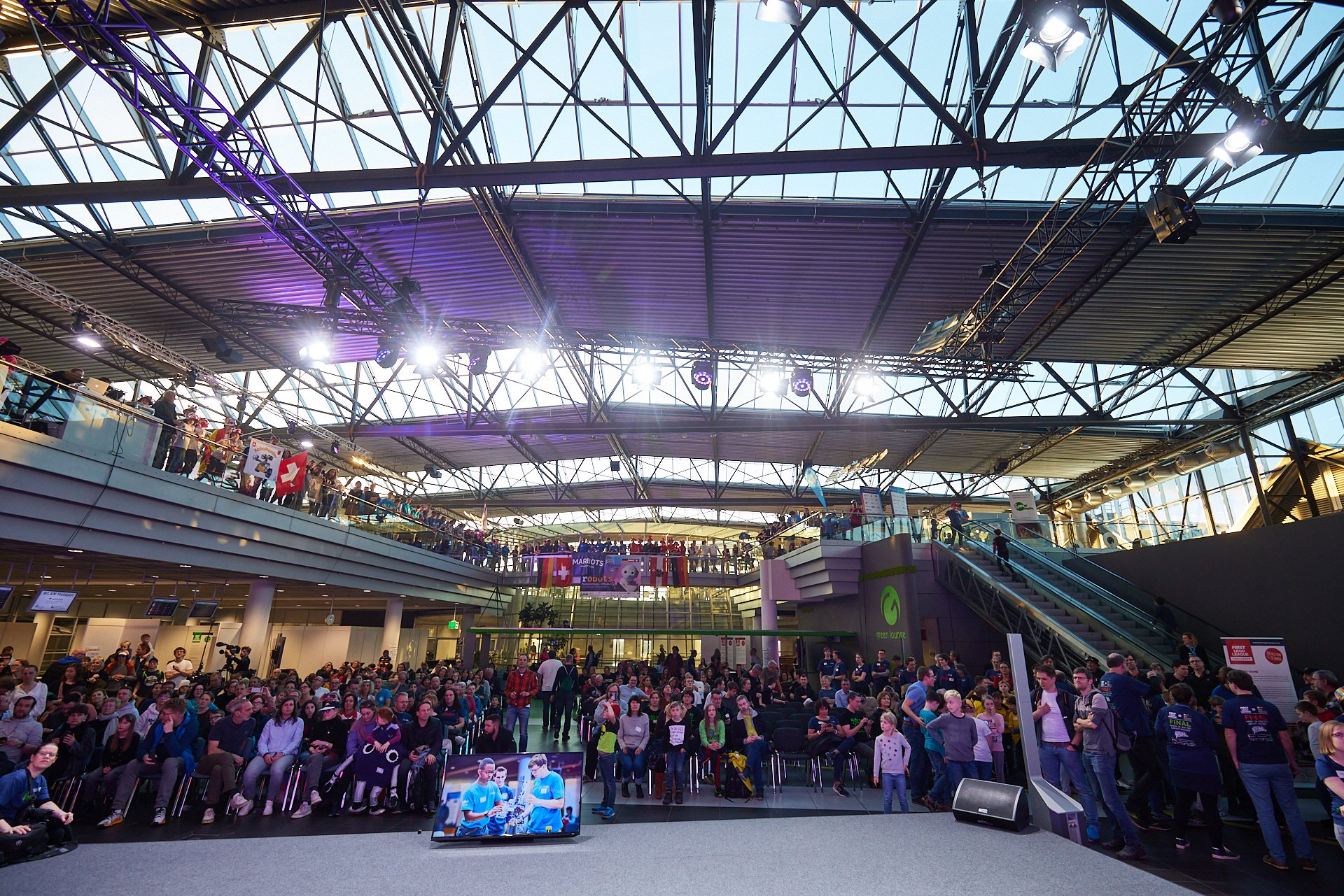 Innenansicht eines geschäftigen Veranstaltungsortes mit Menschen, die auf einer Bühne sitzen und zuschauen, während im Hintergrund mehrere Etagen mit Zuschauern gefüllt sind, under einem großen Glasdach.