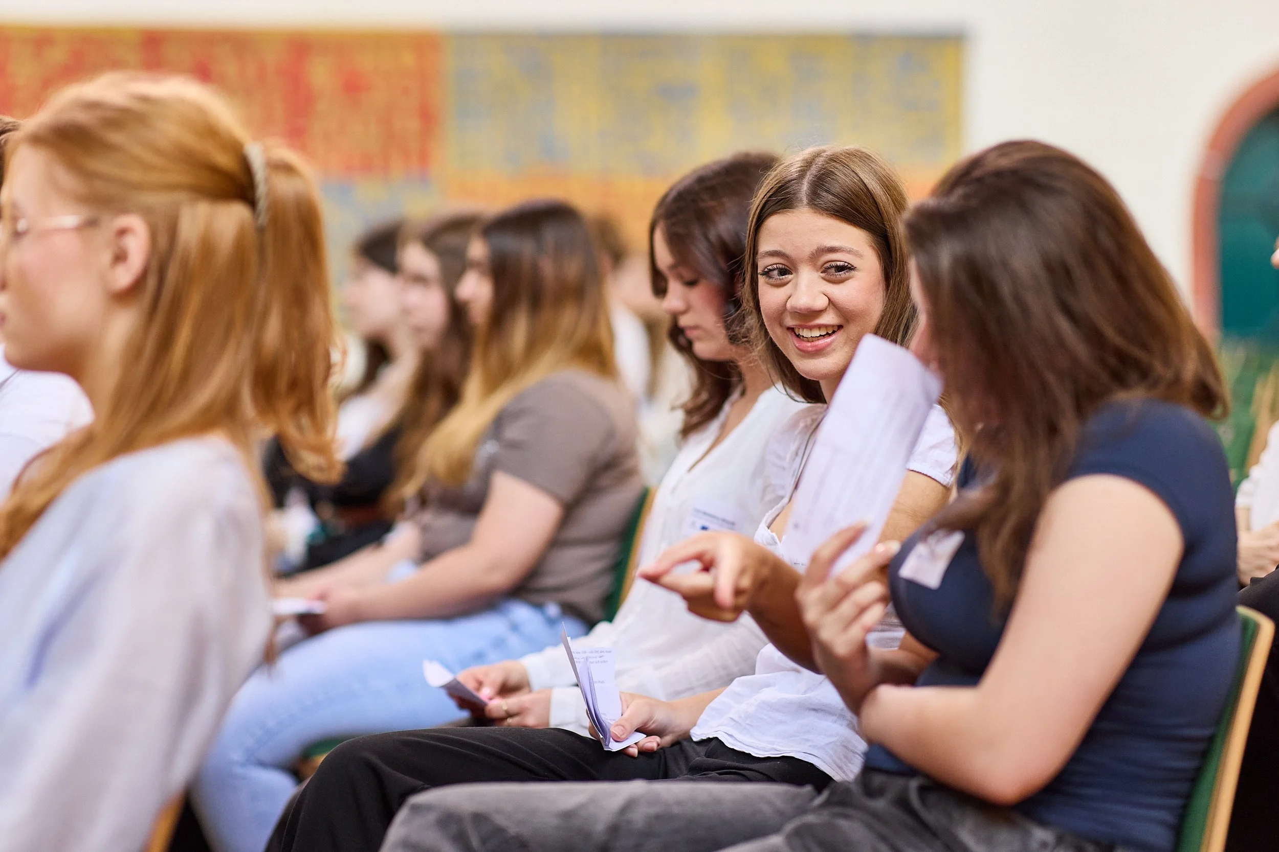 Junge Frau mit braunen Haaren und weißem Hemd lächelt in die Kamera, während sie neben einer anderen Frau sitzt, die eine Pinnwand oder Präsentation betrachtet, bei einer Versammlung oder Schulveranstaltung.