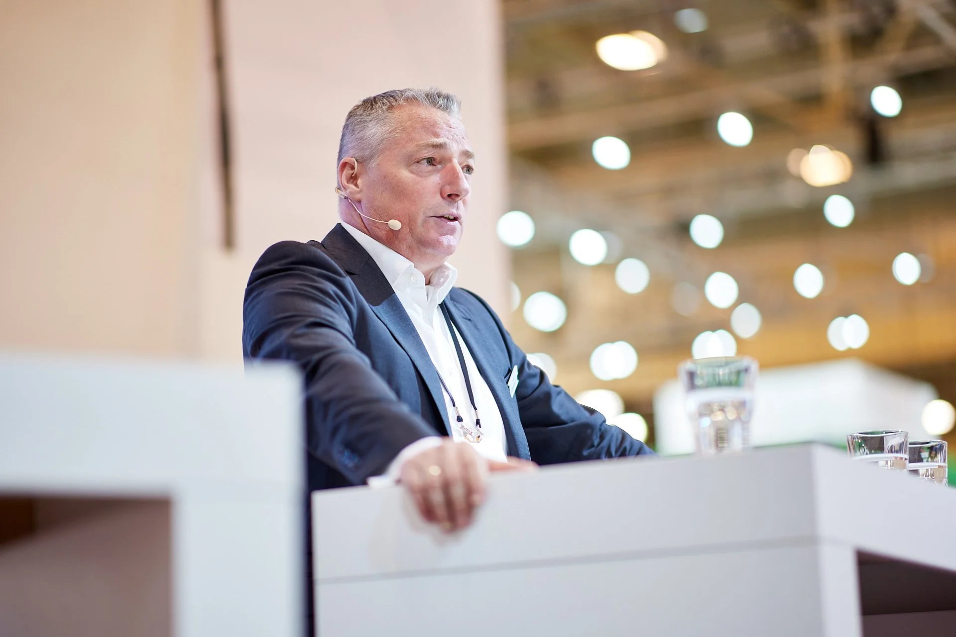 Ein Mann spricht während einer Konferenz, trägt Anzug und Hemd, mit einem Headset-Mikrofon, im Hintergrund viele Lichter.