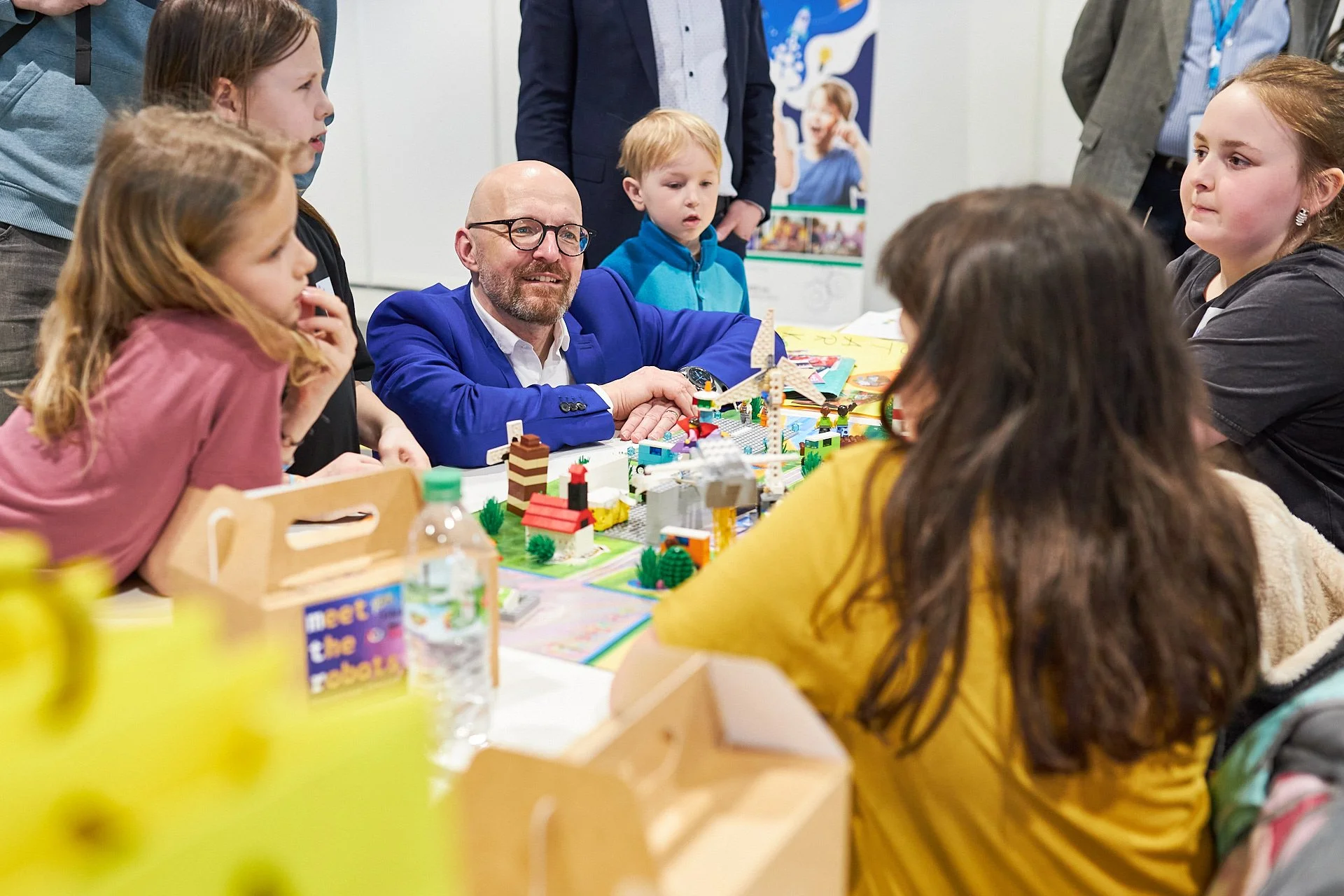 Kinder und Erwachsene sitzen um einen Tisch, der mit LEGO-Bausätzen und Bausteinen dekoriert ist, bei einer Veranstaltung.