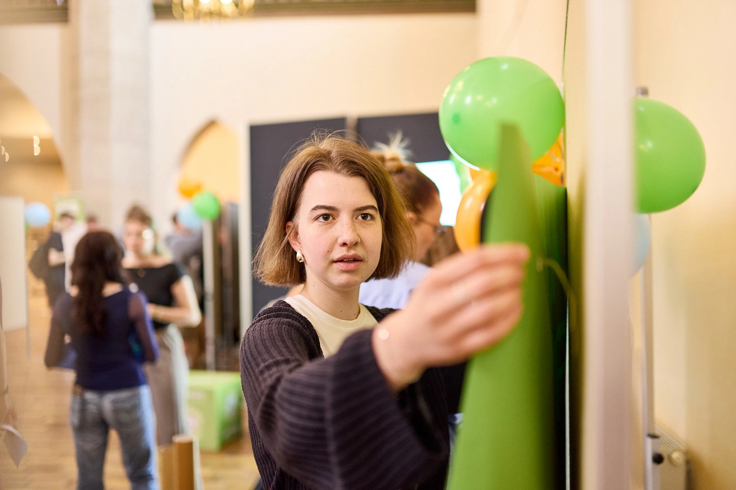 Junge Frau, die an einer Wand mit grünen Ballons befestigt ist, bei einer Veranstaltung mit mehreren Menschen im Hintergrund.