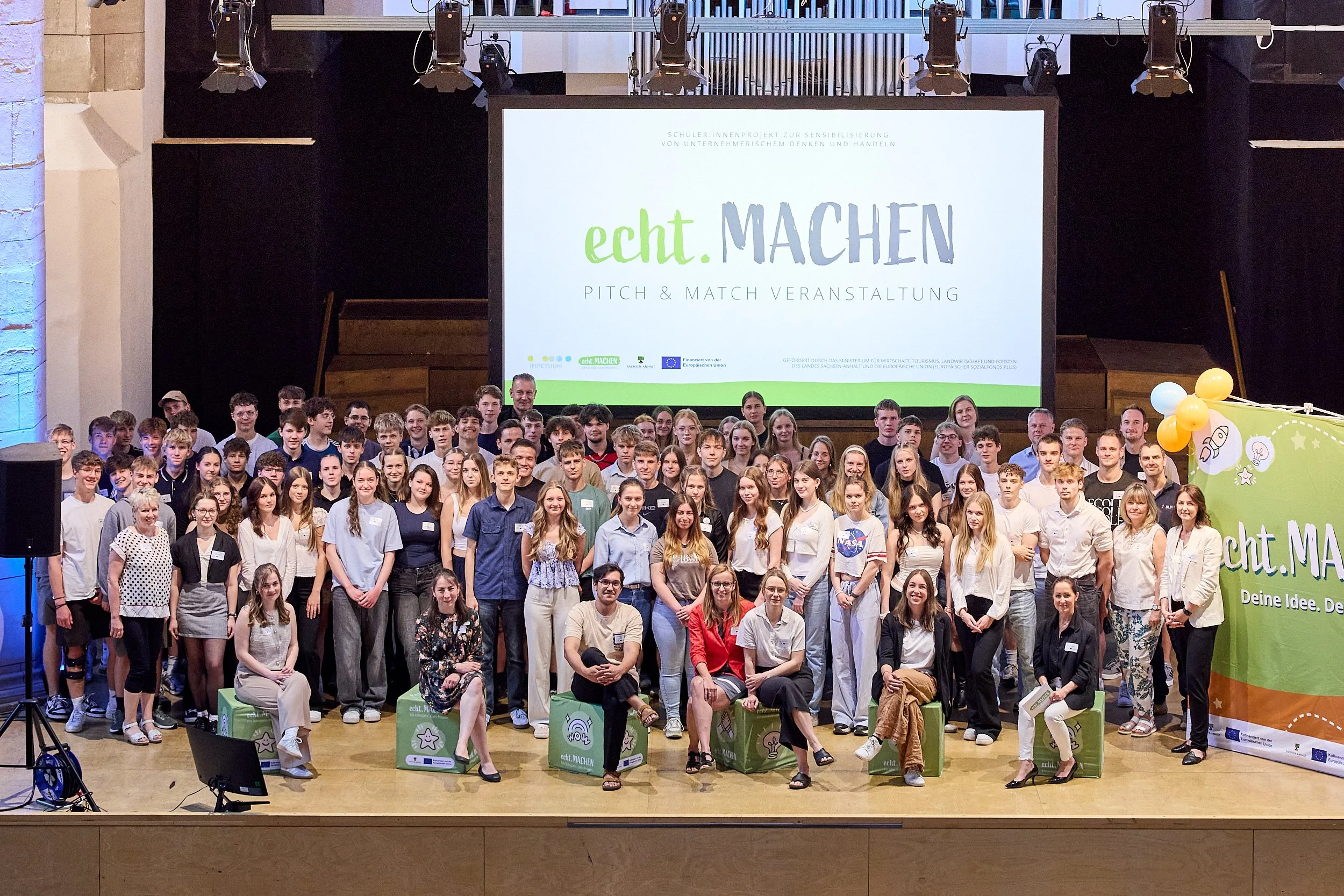 Ein großer Gruppenfoto von Jugendlichen und Erwachsenen auf einer Bühne bei einer Veranstaltung namens 'echt. MACHE' mit einem großen Bildschirm im Hintergrund, auf dem der Veranstaltungstitel angezeigt wird.
