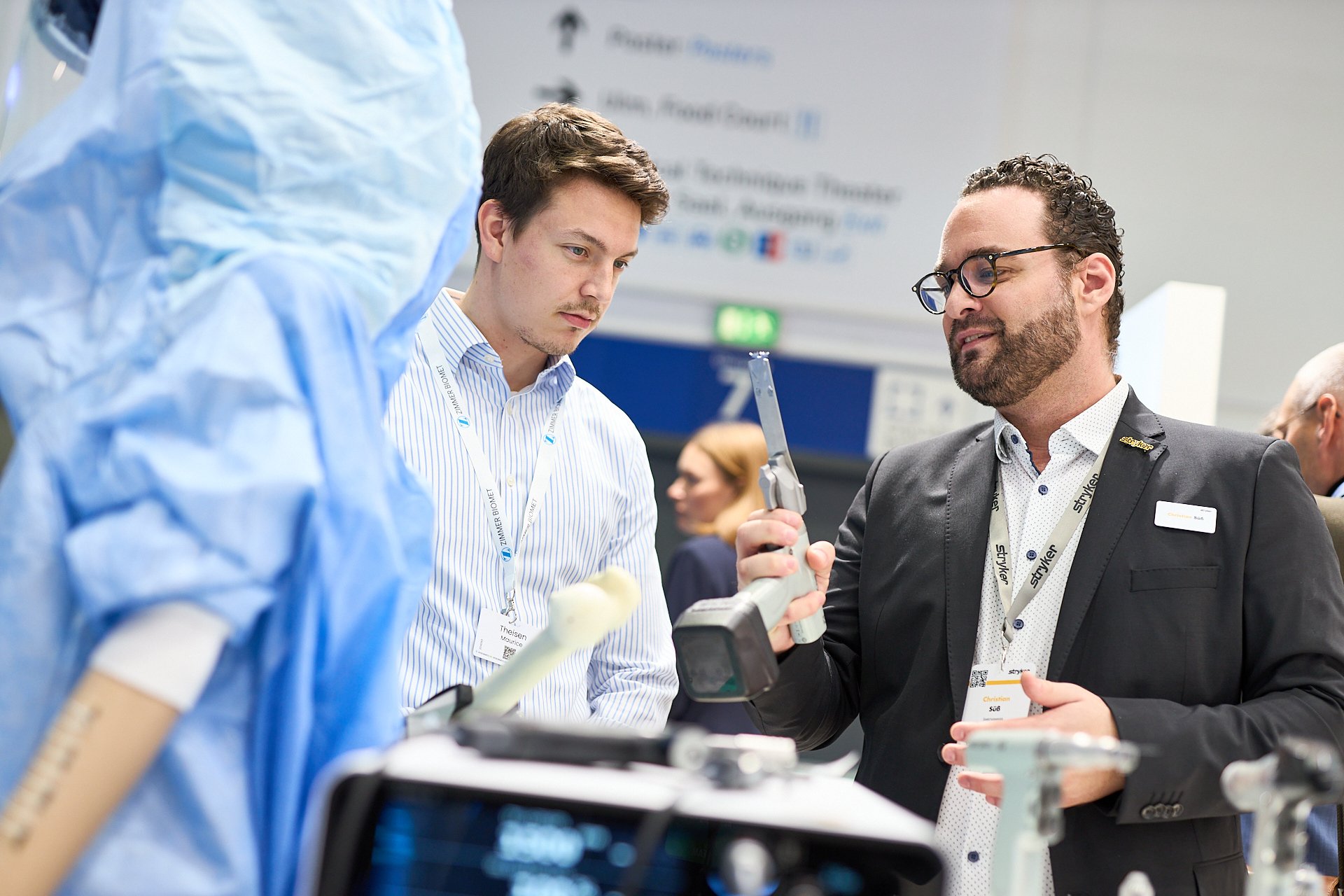 Drei Menschen in einem Gespräch bei einer medizinischen Präsentation, mit medizinischem Equipment im Vordergrund.