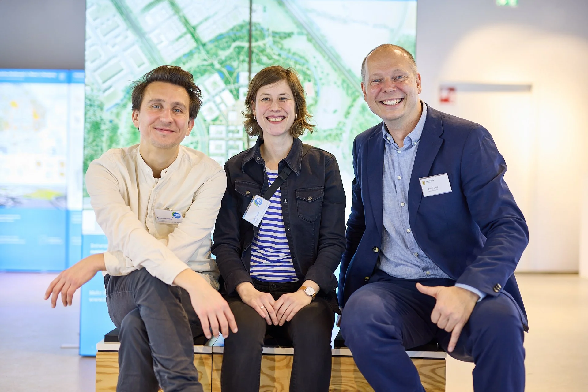 Drei Menschen, zwei Männer und eine Frau, sitzen auf einer Bank in einem modernen Gebäude, lachen und posieren für die Kamera. Hinter ihnen ist eine große digitale Karte oder einen Stadtplan sichtbar.