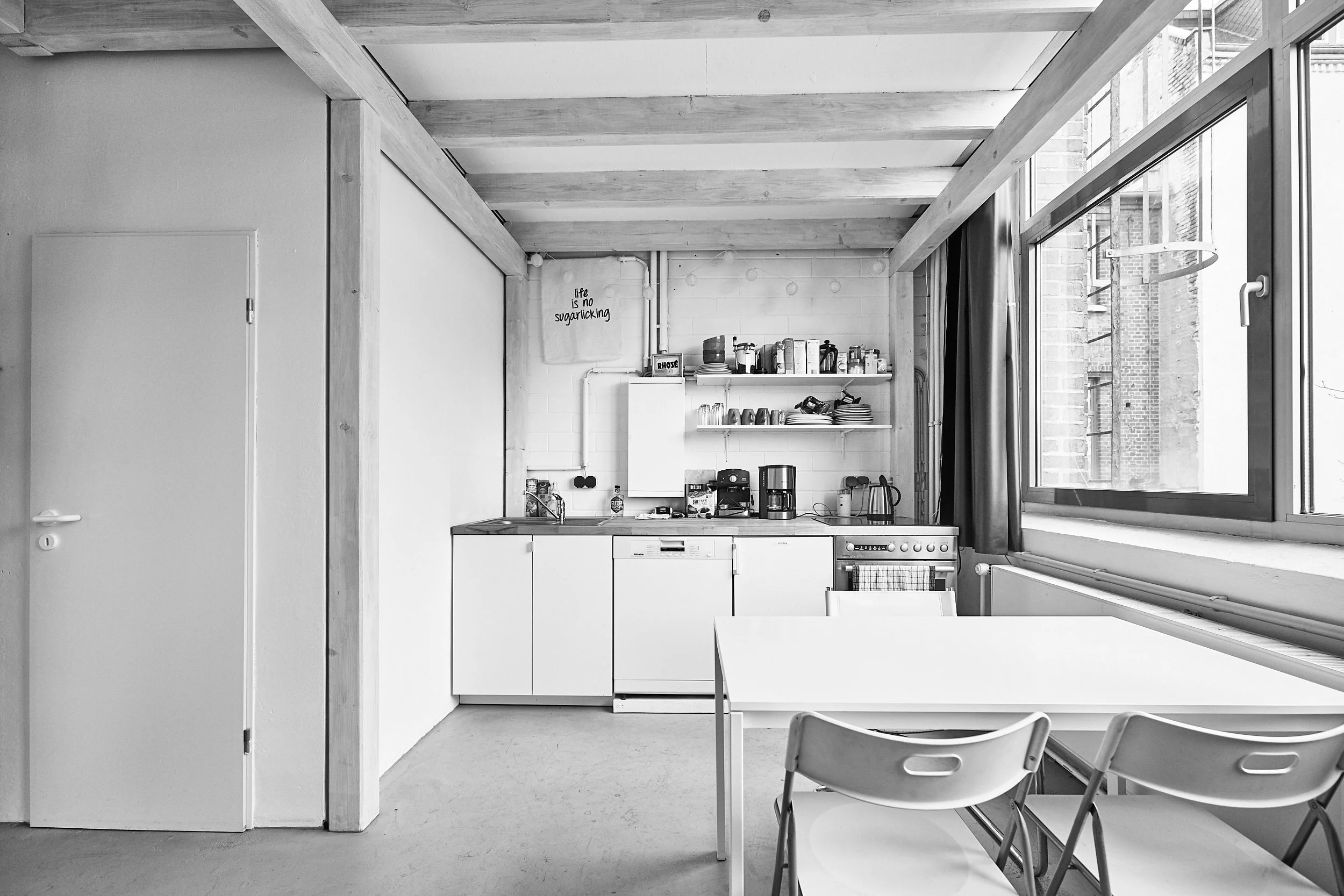 Ein minimalistisches, schwarz-weißes Kücheninterieur mit weißen Schränken, einem Esszimmertisch und Stühlen, einem Fenster auf der rechten Seite und offenen Regalen mit Küchenutensilien.