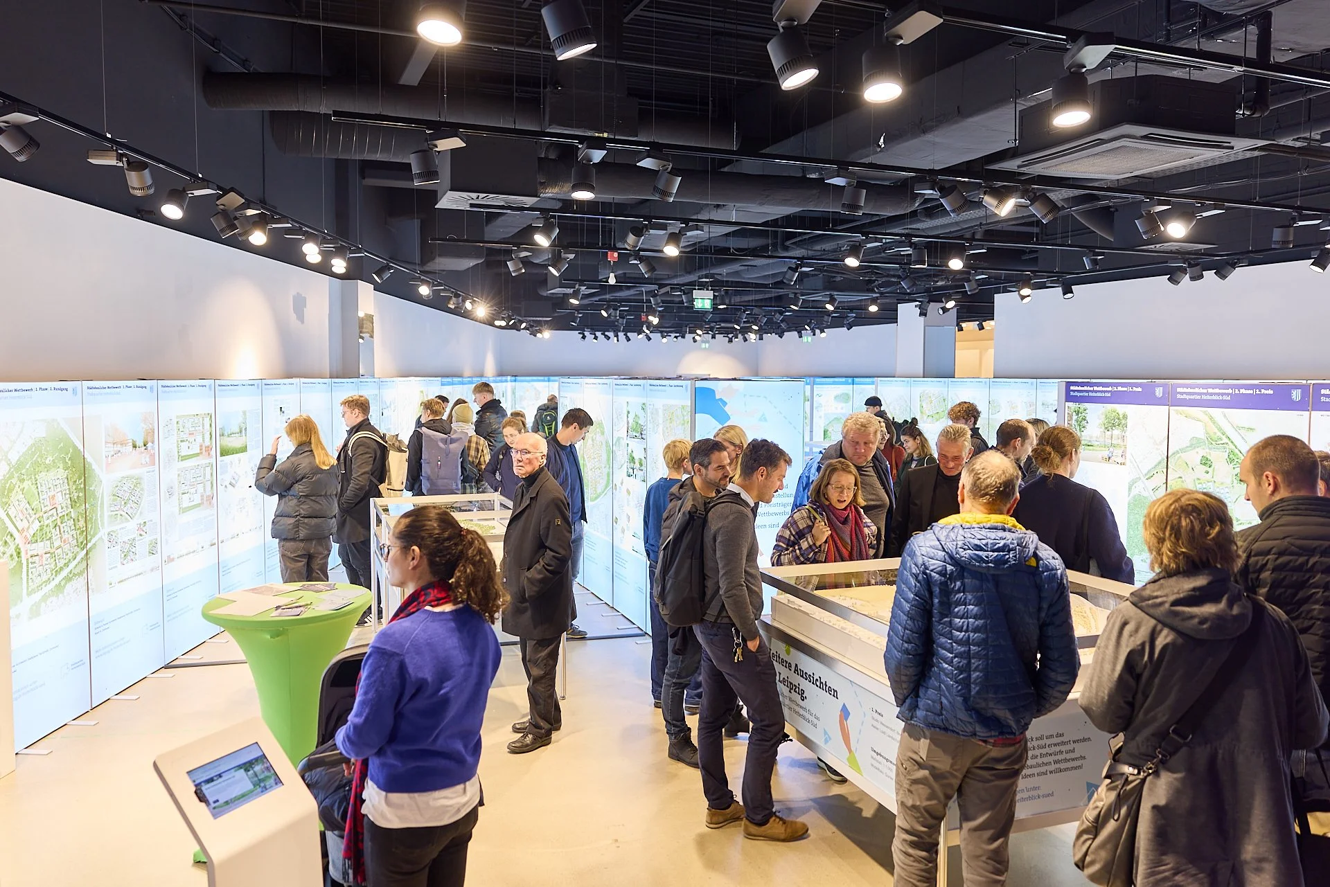 Menschen betrachten informierende Ausstellung mit großen Bildtafeln und Infotafeln in einem gut beleuchteten, modernen Raum.