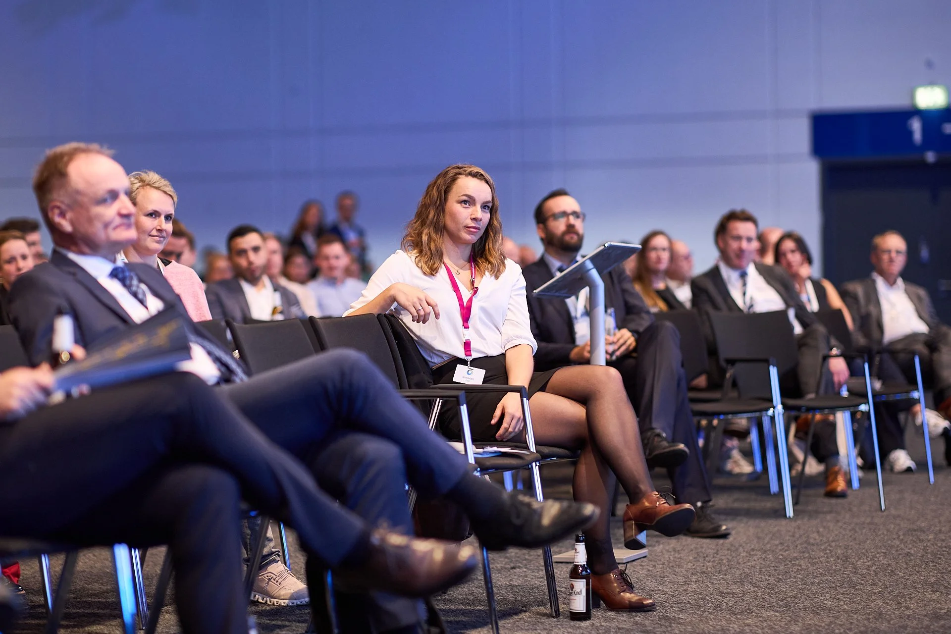 Personen sitzen in einem Seminarraum bei einer Konferenz, manche nehmen Notizen, andere hören aufmerksam zu.