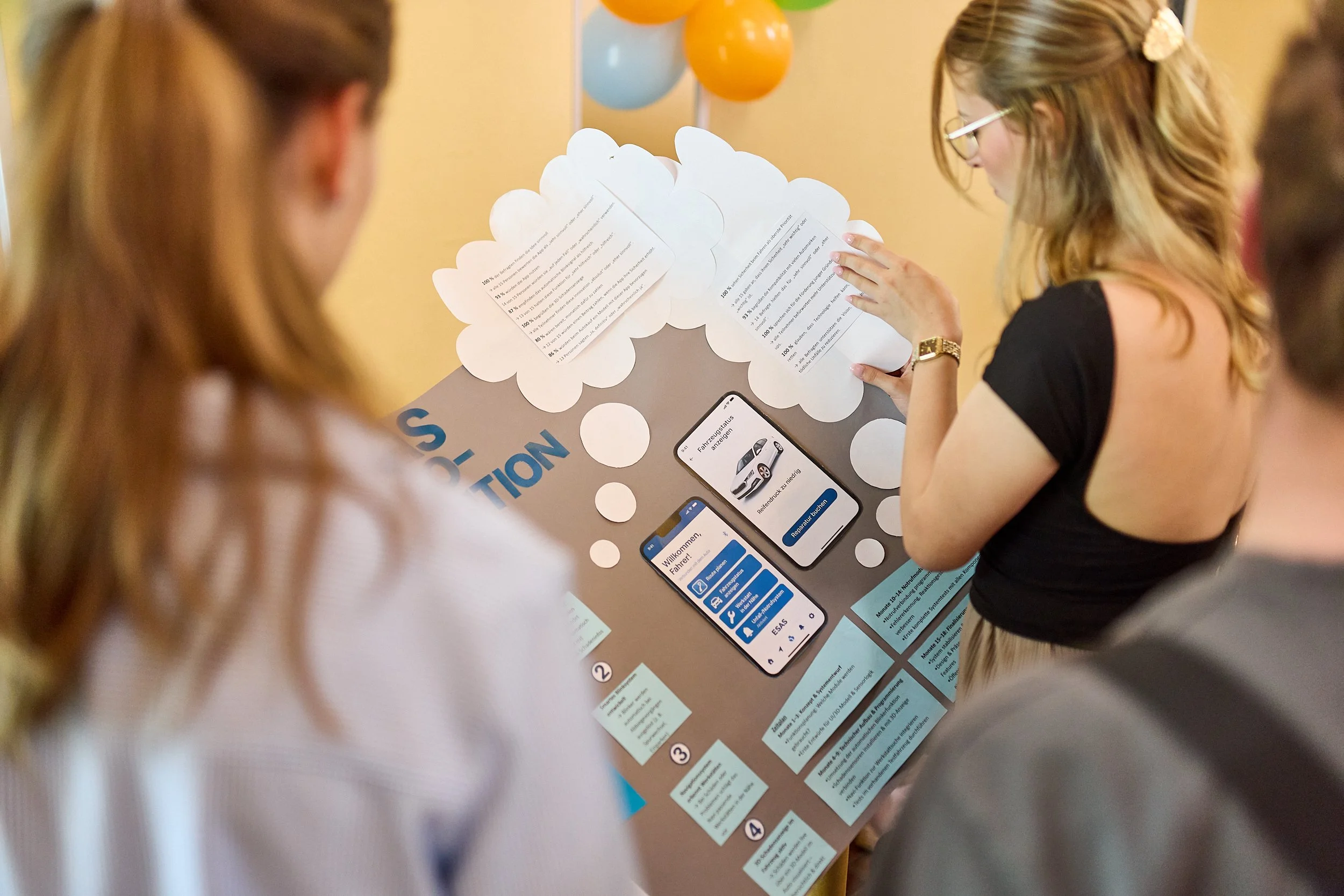 Mehrere Menschen betrachten eine Präsentations- und Informationswand mit Postern, einem Smartphone und Ballons im Hintergrund bei einer Veranstaltung.