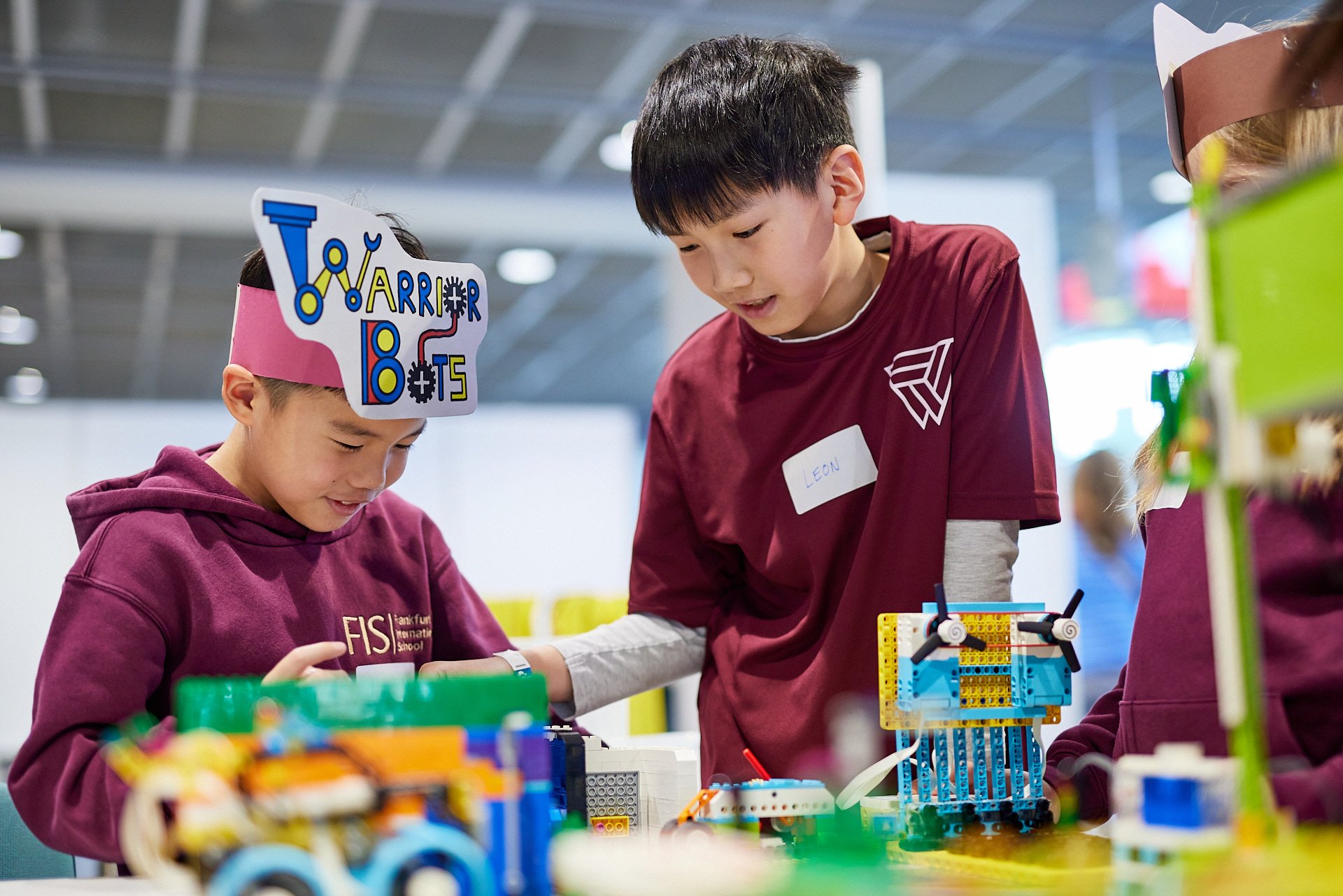 Drei Kinder, die an einem Tisch mit LEGO-Modelle bauen, in einem hellen Raum mit hoher Decke, einem Kind trägt eine Kopfhalterung mit der Aufschrift "Lego Arrier 2005", ein anderes trägt ein Namensschild mit "LEON".