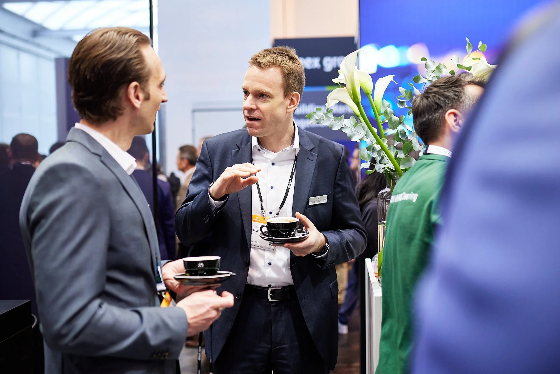 Drei Männer im Gespräch auf einer Konferenz, zwei halten Kaffeetassen, im Hintergrund andere Teilnehmer und ein großer Bildschirm.