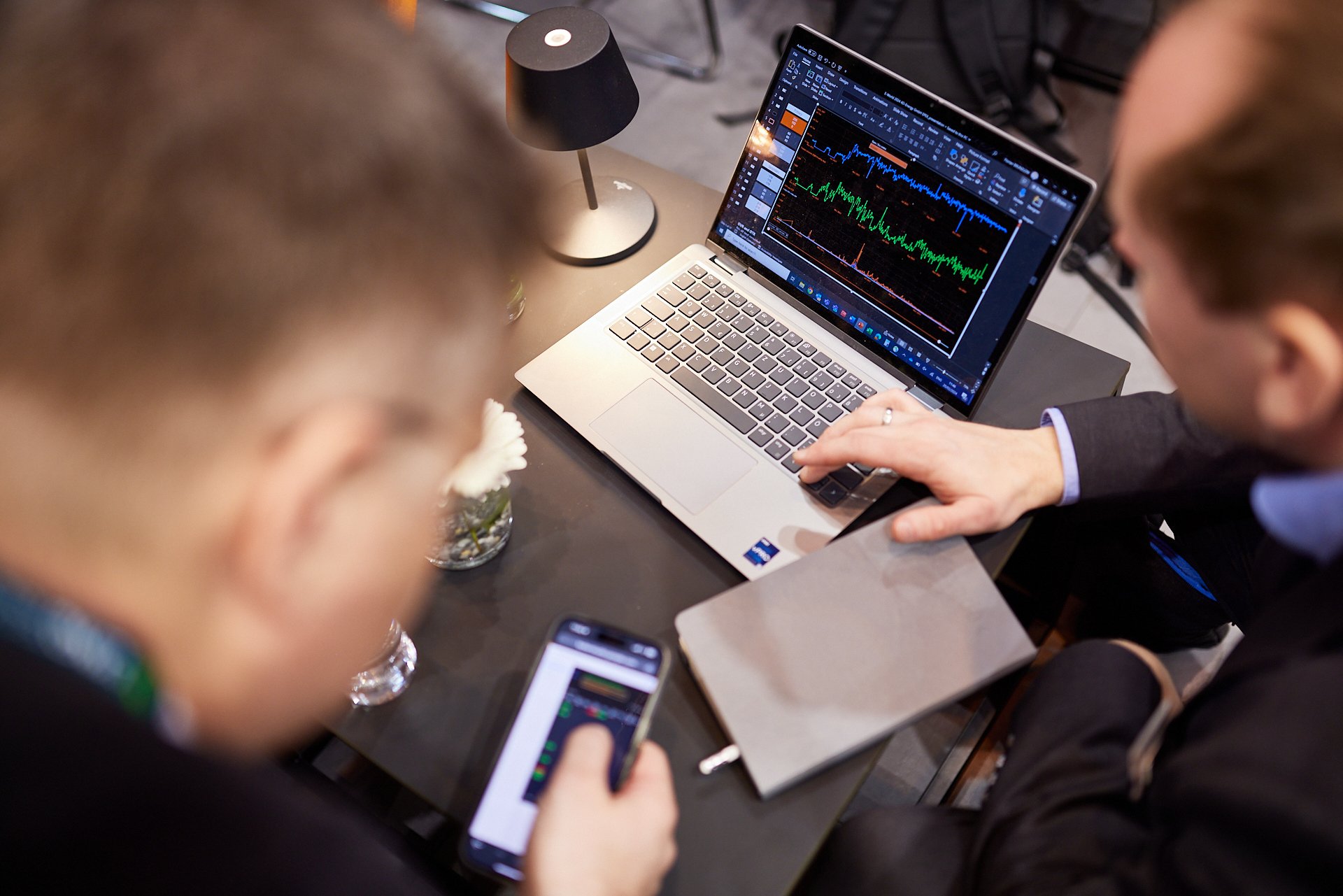 Zwei Personen beobachten Grafiken auf einem Laptop, einer nutzt ein Smartphone, ein Notizbuch liegt auf dem Tisch, am Tisch steht eine kleine Tischlampe.