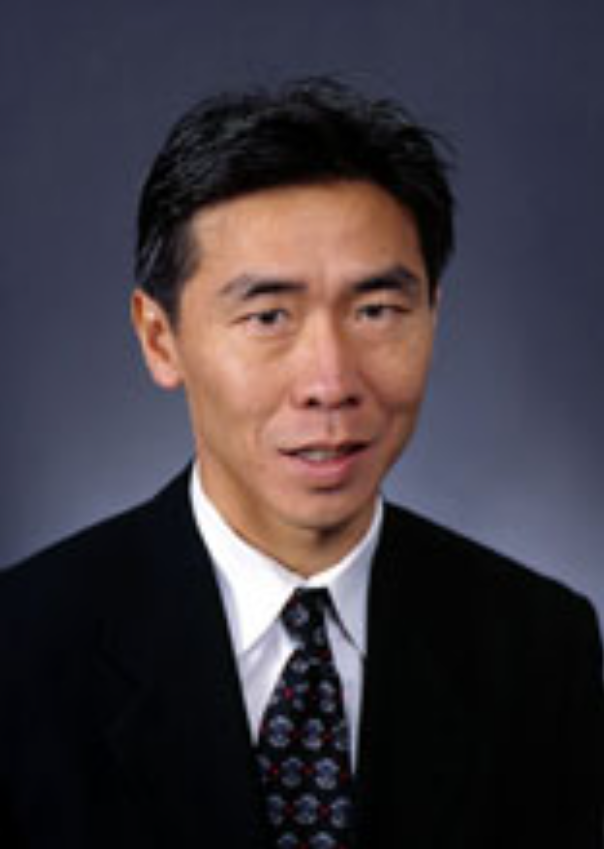 A man in a black suit with a white shirt and patterned tie, against a dark background.