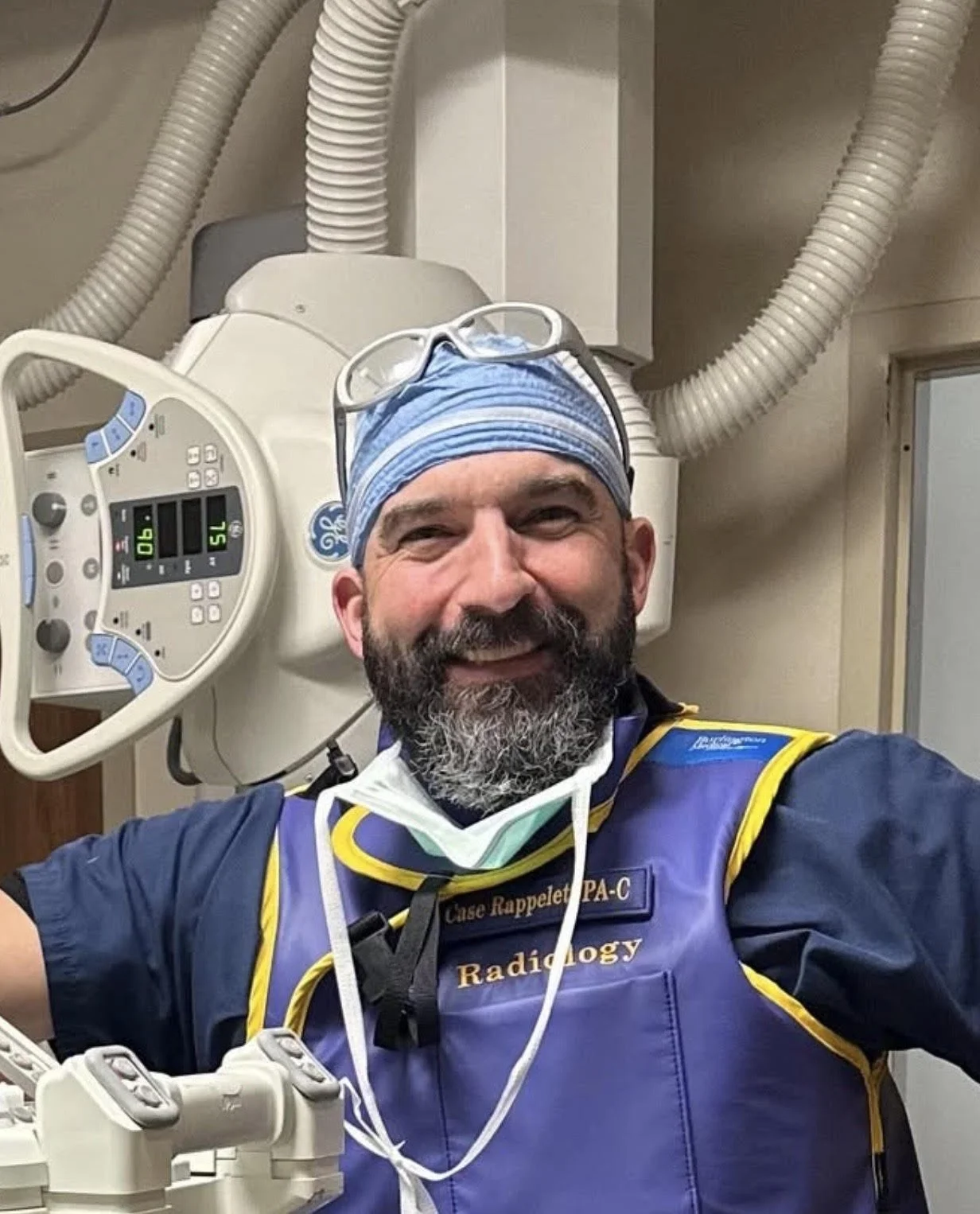 A smiling man in medical scrubs and a surgical cap with glasses resting on his head, standing in a hospital or medical setting with medical equipment behind him.