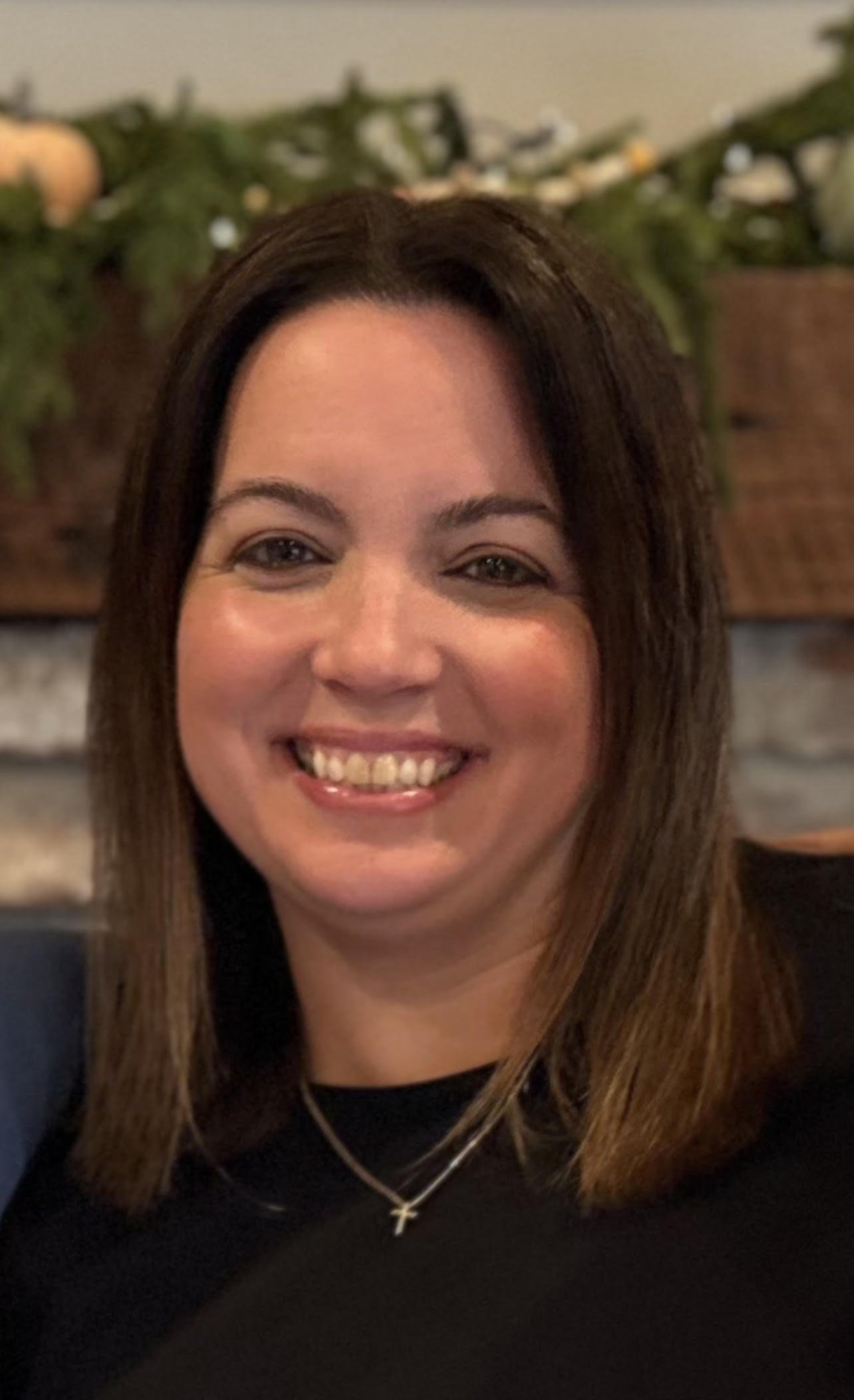 A smiling woman with brown hair and a cross necklace, in front of a brick wall decorated with greenery and ornaments.