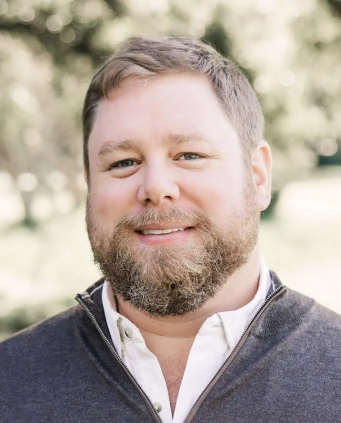 A man with light brown hair, beard, and blue eyes, wearing a white lab coat and a checkered shirt, smiling in front of a plain white background.