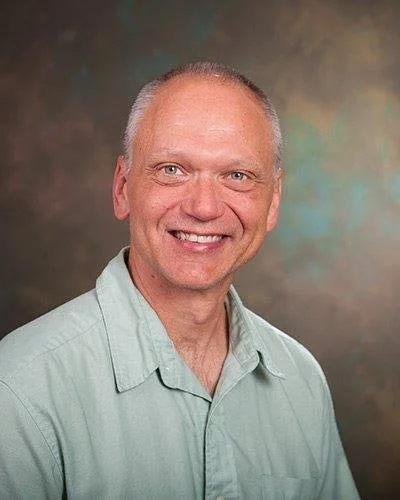 A smiling man with short gray hair wearing a light-colored collared shirt.