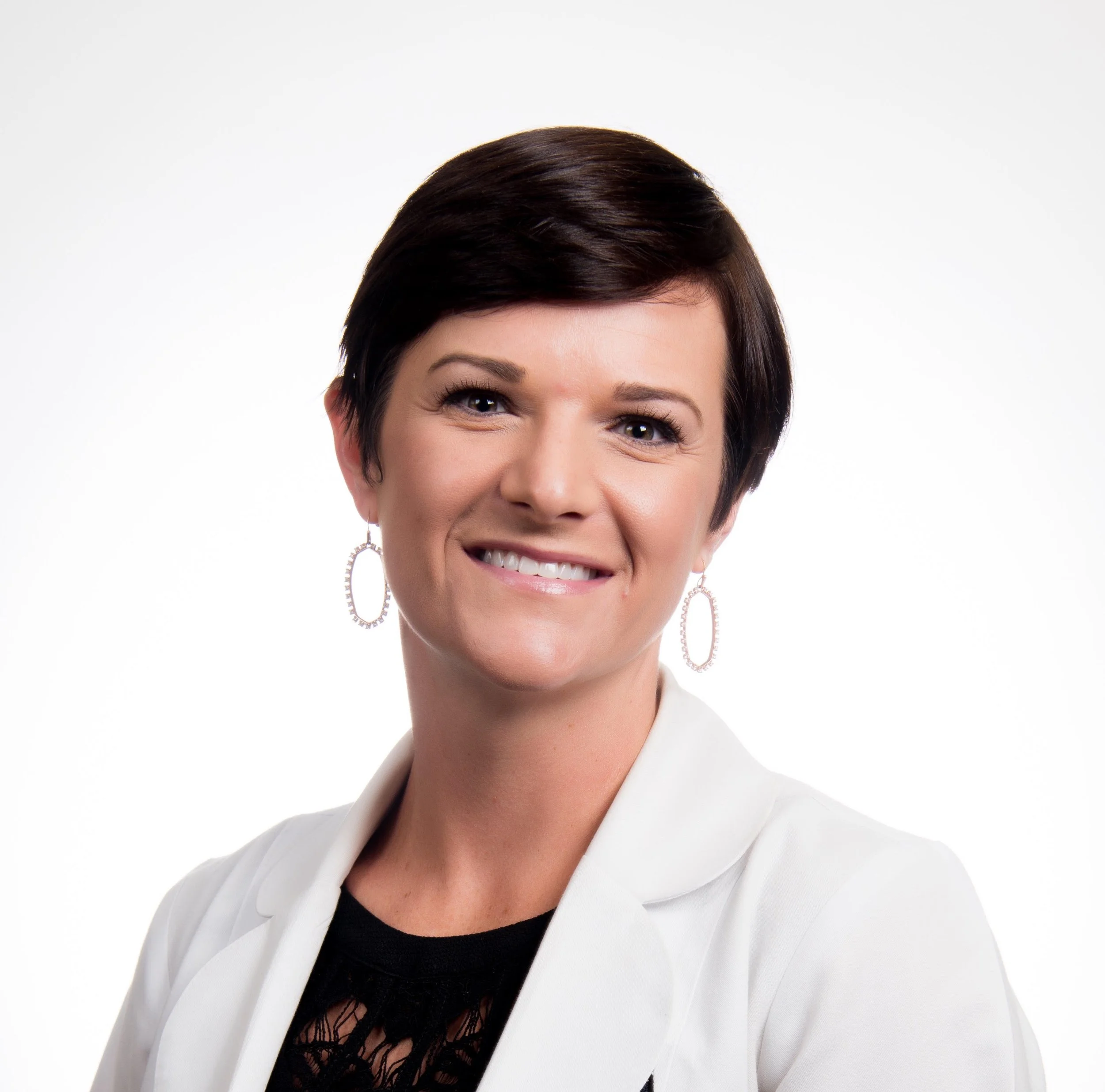 Professional woman with short dark hair, smiling, wearing earrings and a white blazer against a plain background.