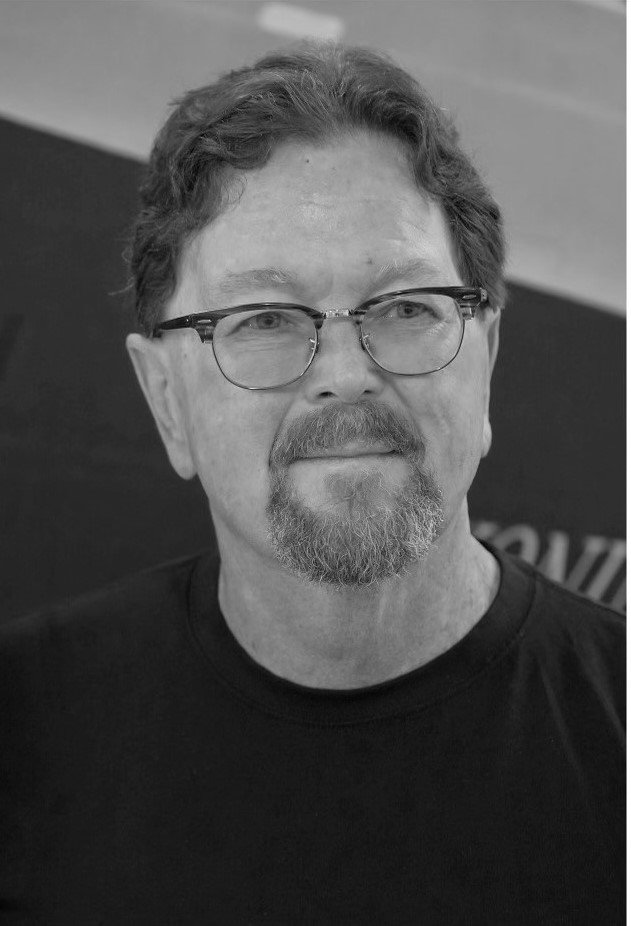 Black and white photo of a man with glasses, a beard, and short wavy hair, wearing a black shirt, smiling slightly.