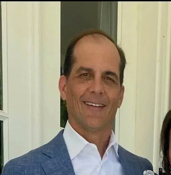 A man in a dark suit with a blue shirt and tie, smiling, against a plain background.