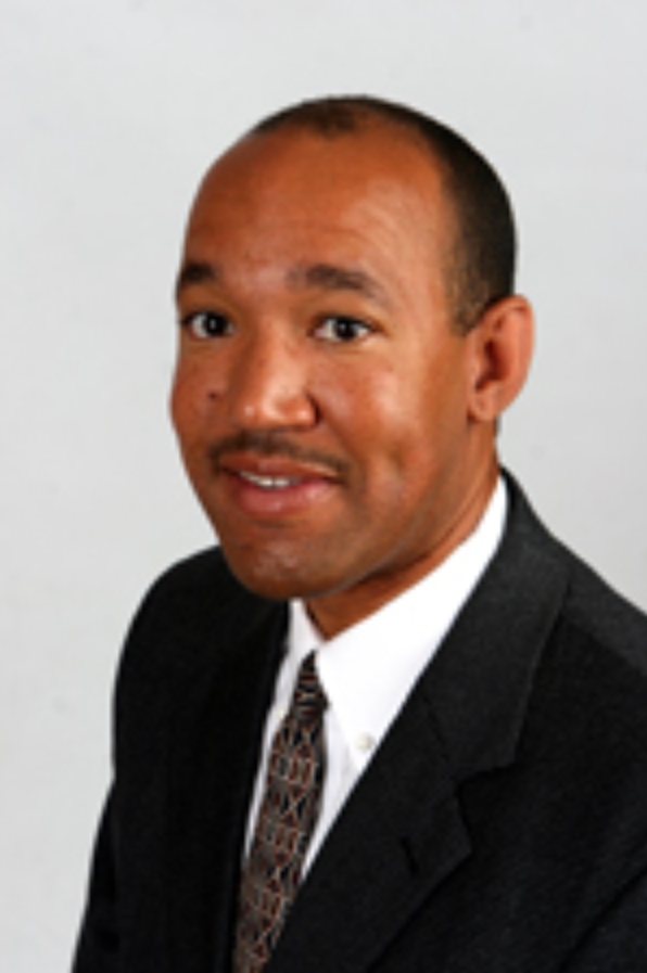 A man in a suit and tie smiling at the camera, with a plain light background.