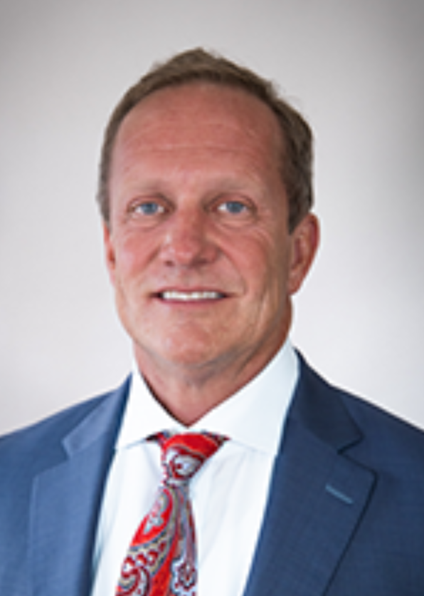 Portrait of a middle-aged man with blue eyes and blonde hair, wearing a dark blue suit, white shirt, and red patterned tie, against a plain background.