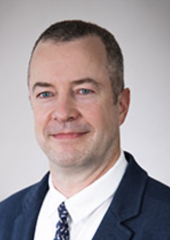 Portrait of a man in a business suit with a white shirt and patterned tie, smiling, with a plain light gray background.