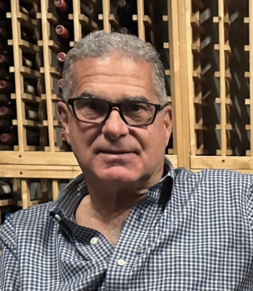 A man with short gray hair and glasses, wearing a dark shirt, standing against a plain background.