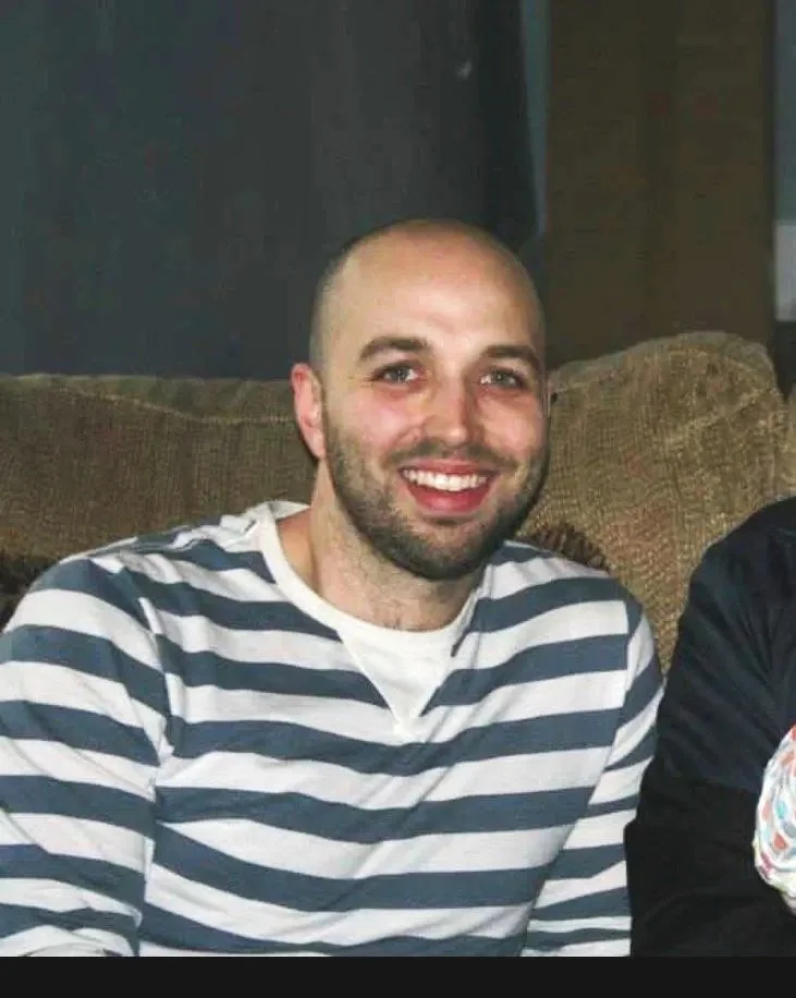 A middle-aged man sitting on a bench, wearing a brown polo shirt and white pants, looking thoughtfully to the side against a plain wall.
